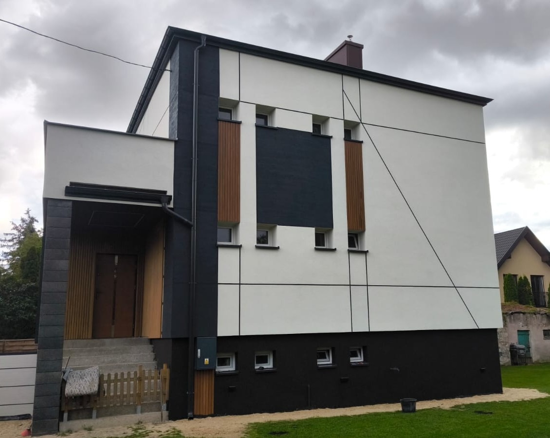 Nowoczesna elewacja domu jednorodzinnego z elementami dekoracyjnymi w kolorach bieli, czerni i drewna, widok z zewnątrz, w tle pochmurne niebo.