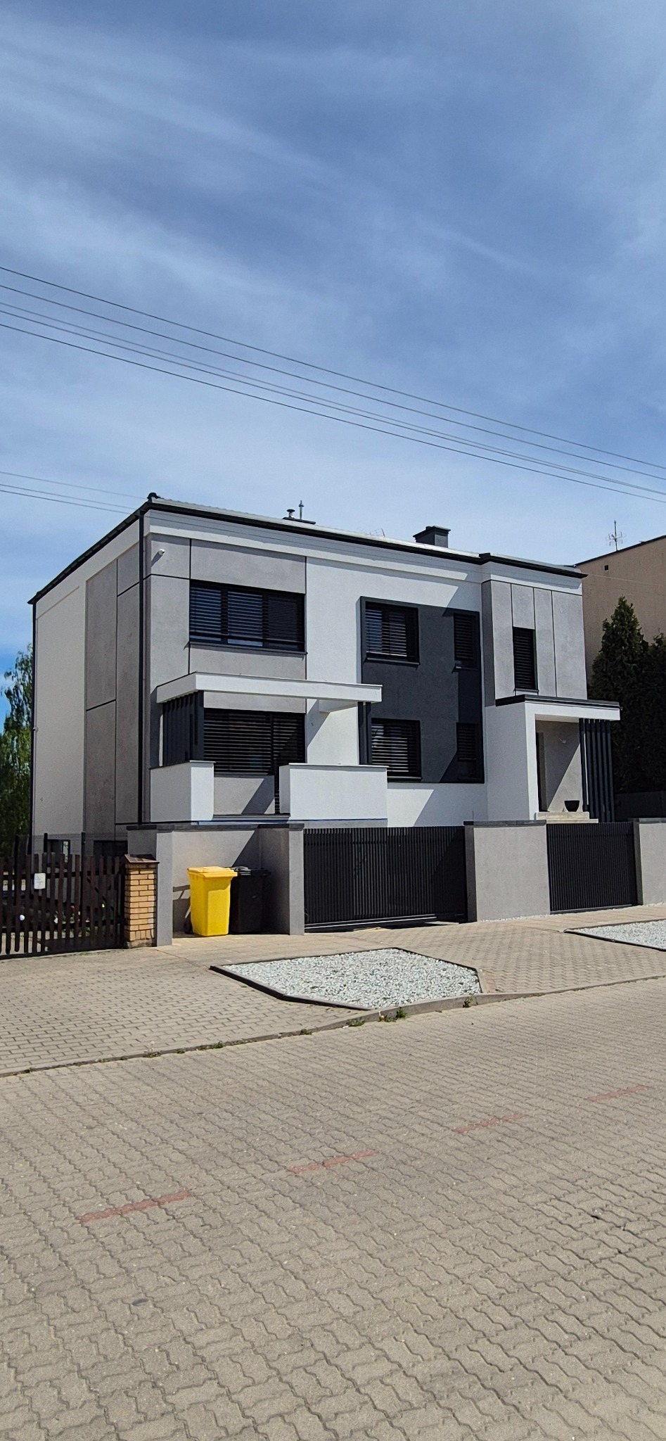 Dwukondygnacyjny dom z elewacją w odcieniach szarości i bieli, czarnymi żaluzjami okiennymi i żółtym pojemnikiem na śmieci przed posesją.