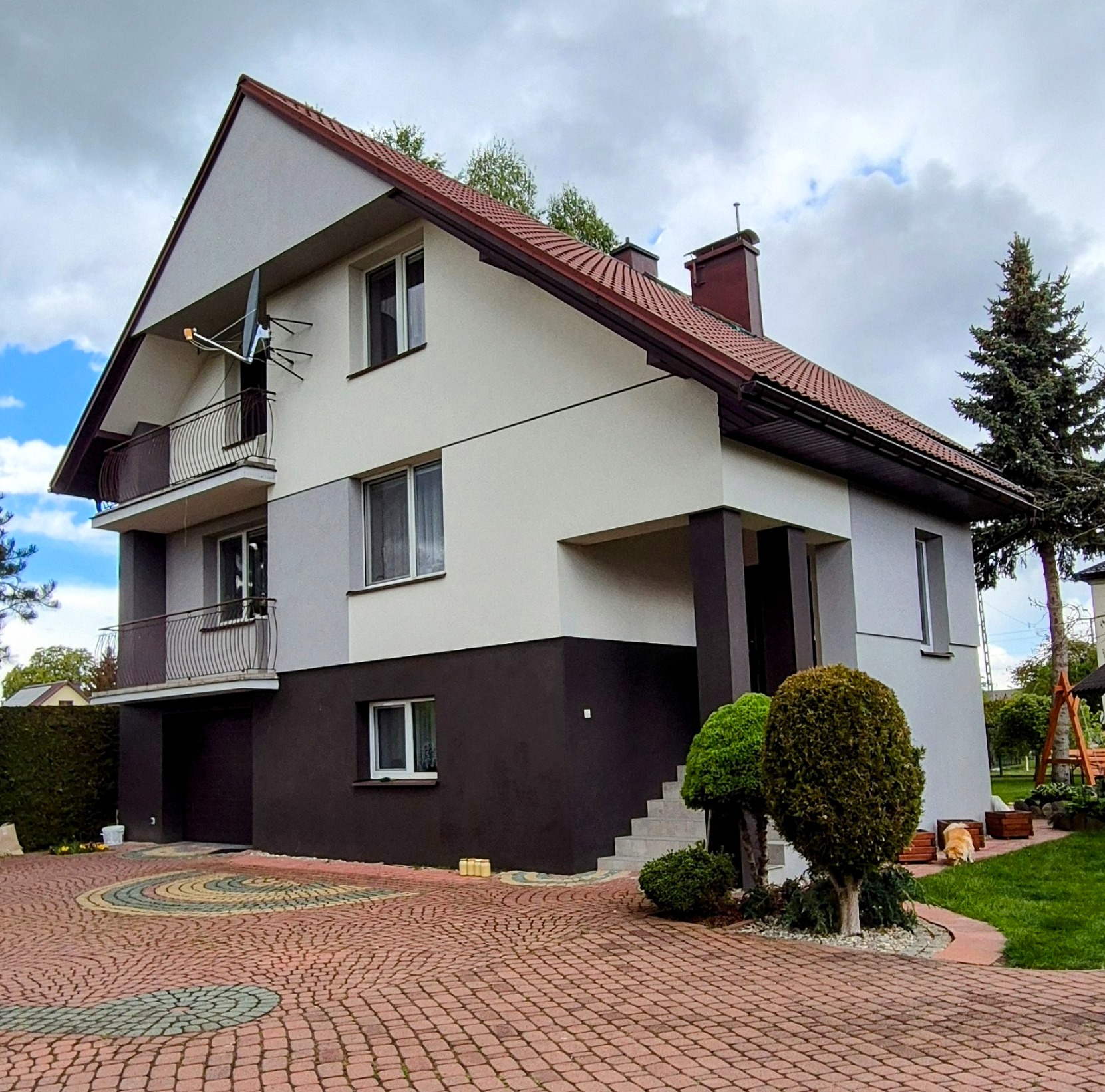 Dwukondygnacyjny dom z balkonami, pomalowany w odcieniach szarości i brązu, z widoczną anteną satelitarną na elewacji i ozdobnym brukiem na podjeździe.