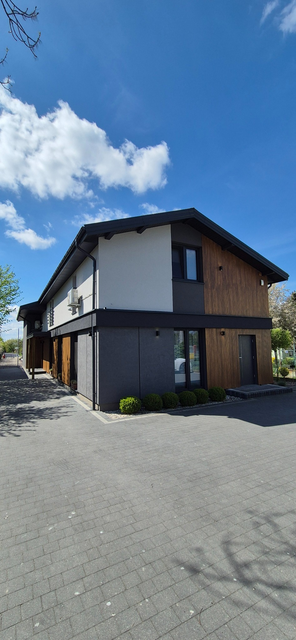 Nowoczesny budynek z elewacją w odcieniach szarości, bieli i drewna. Widoczne klimatyzatory, małe krzewy i chodnik z kostki brukowej. Błękitne niebo z chmurami w tle.