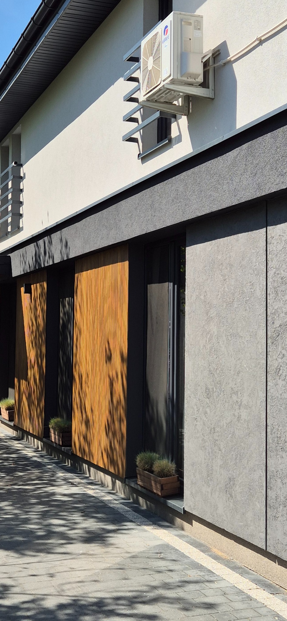 Nowoczesna elewacja budynku z drewnianymi panelami i klimatyzatorem, oświetlona naturalnym światłem. Widoczne rośliny w donicach i cień drzewa.