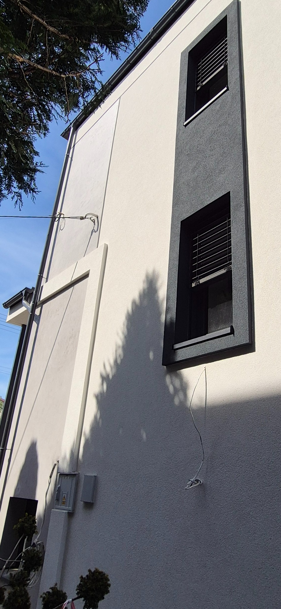 Wykończona elewacja domu z oknami w grafitowych obramowaniach, widok z dołu, Białystok. Jasna fasada z elementami dekoracyjnymi i cieniem drzewa.