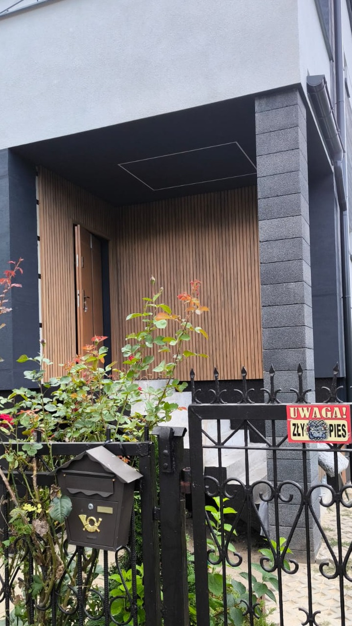 Nowoczesna elewacja domu z drewnianą okładziną wejściową, czarnymi elementami i szarą fasadą. Ogródek z roślinami i skrzynką pocztową.