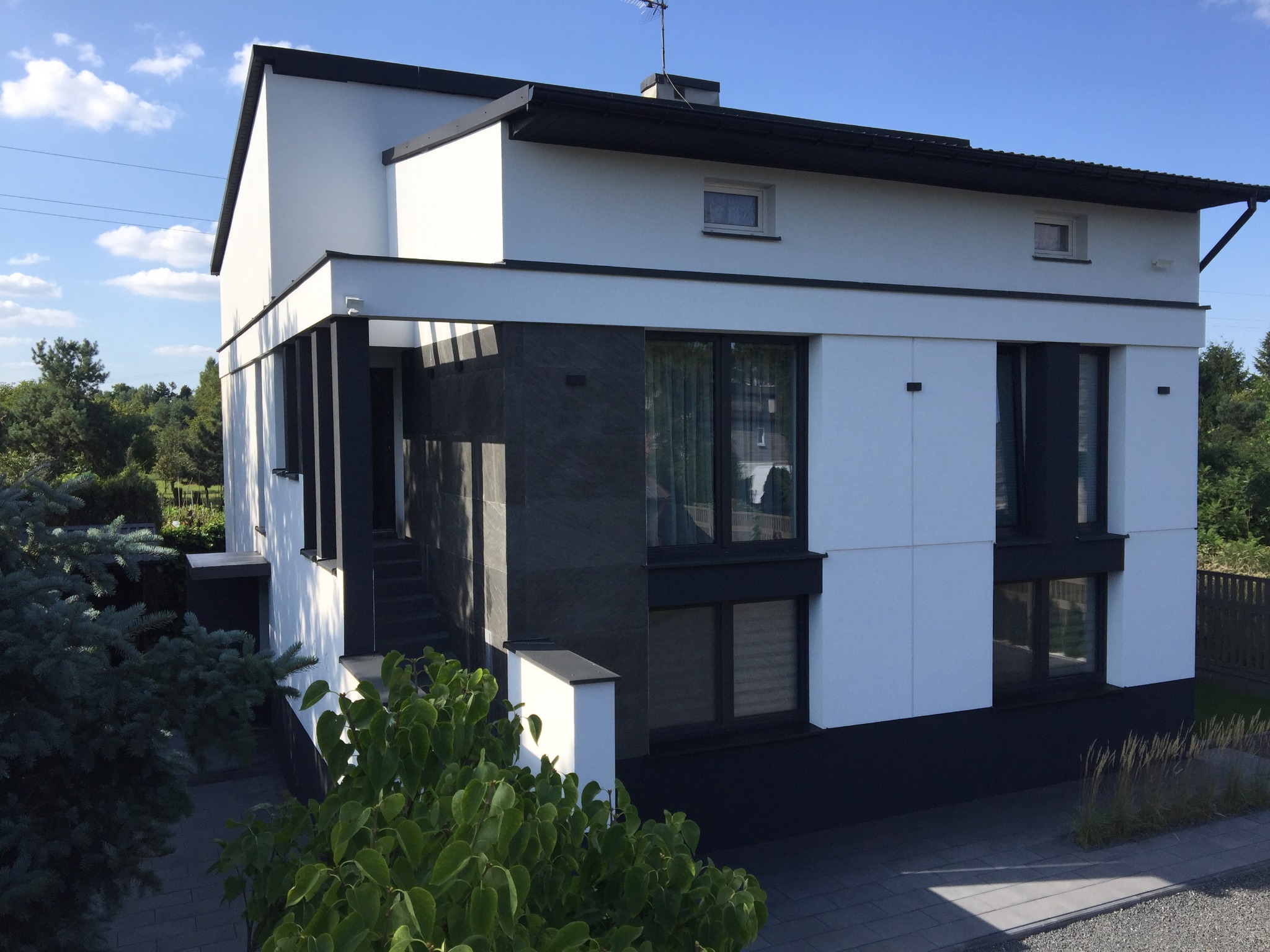 Nowoczesny dom z białą elewacją i czarnymi akcentami, widok z zewnątrz w słoneczny dzień. Minimalistyczny design, duże okna, zadbany ogród.