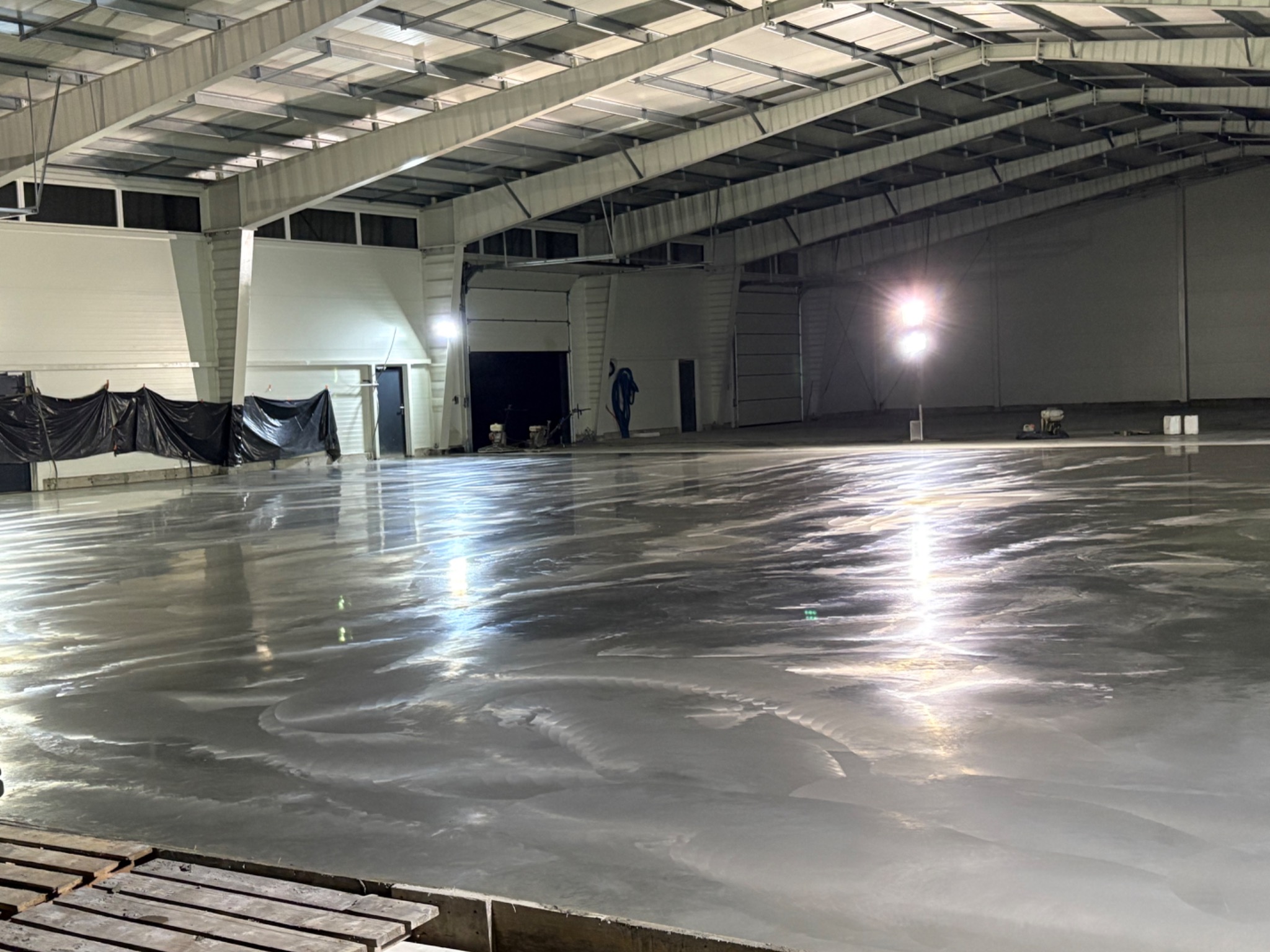 Błyszcząca, świeżo wylana posadzka przemysłowa w hali z widocznymi śladami po maszynach i ochronną folią przy ścianach. Widoczne oświetlenie i konstrukcja dachu.