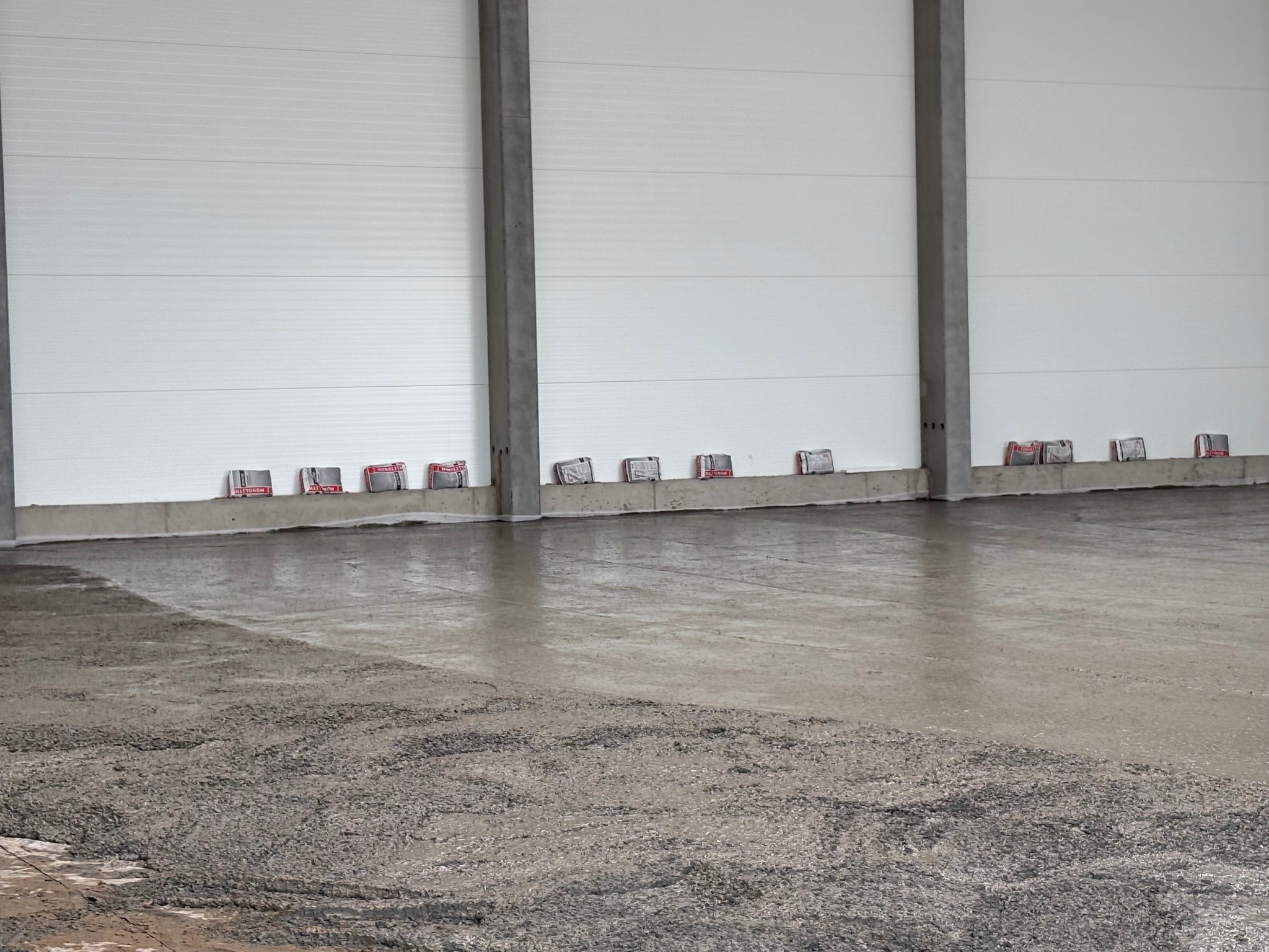 Wylewka betonowa na podłożu hali przemysłowej, widoczne pakiety izolacyjne przy ścianie. Częściowo wykonana, gładka powierzchnia.