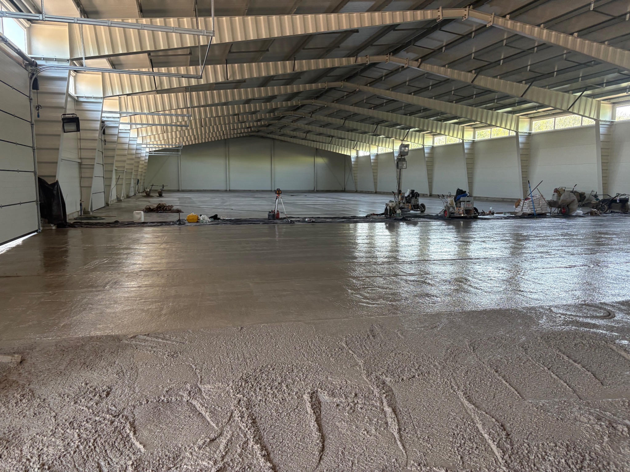 Wnętrze hali z wylewką w trakcie realizacji. Widoczne maszyny do wyrównywania posadzki i ekipa pracująca nad wykończeniem. Częściowo wygładzona powierzchnia w odcieniach szarości.