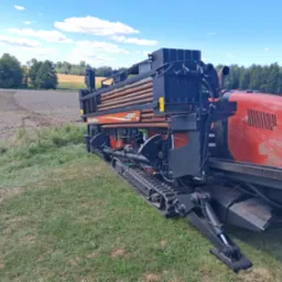 Czerwono-czarna wiertnica Ditch Witch JT2020 Mach1 na gąsienicach, z rurami w zasobniku, na trawiastym terenie z polem w tle, w słoneczny dzień.