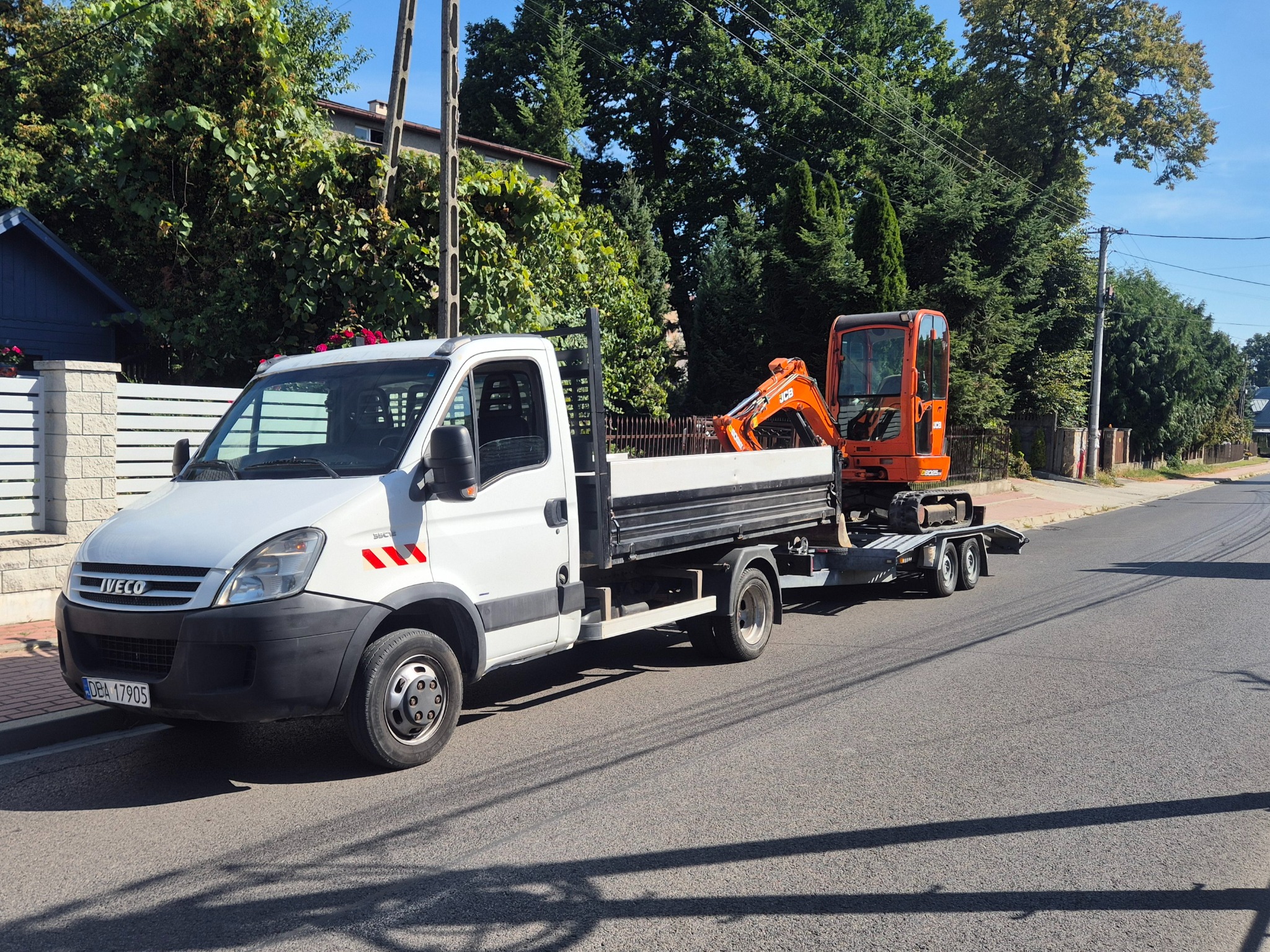 Biały Iveco z pomarańczową mini koparką na lawecie parkuje na ulicy, w tle zieleń drzew i domy. Widoczne tablice rejestracyjne.