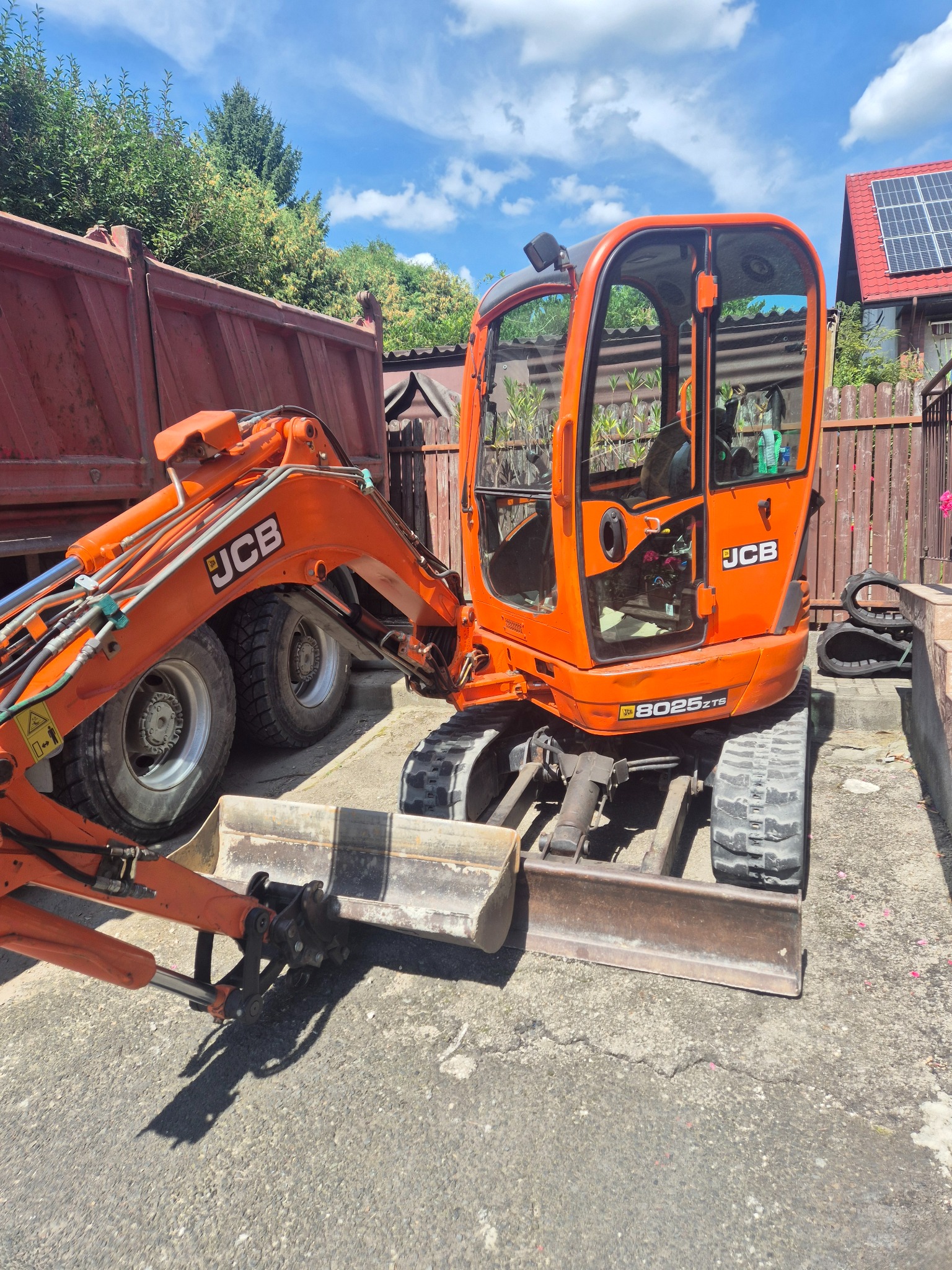 Pomarańczowa mini koparka JCB 8025 ZTS na betonowym podłożu, obok wywrotka. W tle drewniane ogrodzenie i dom z panelami słonecznymi. Słoneczny dzień.