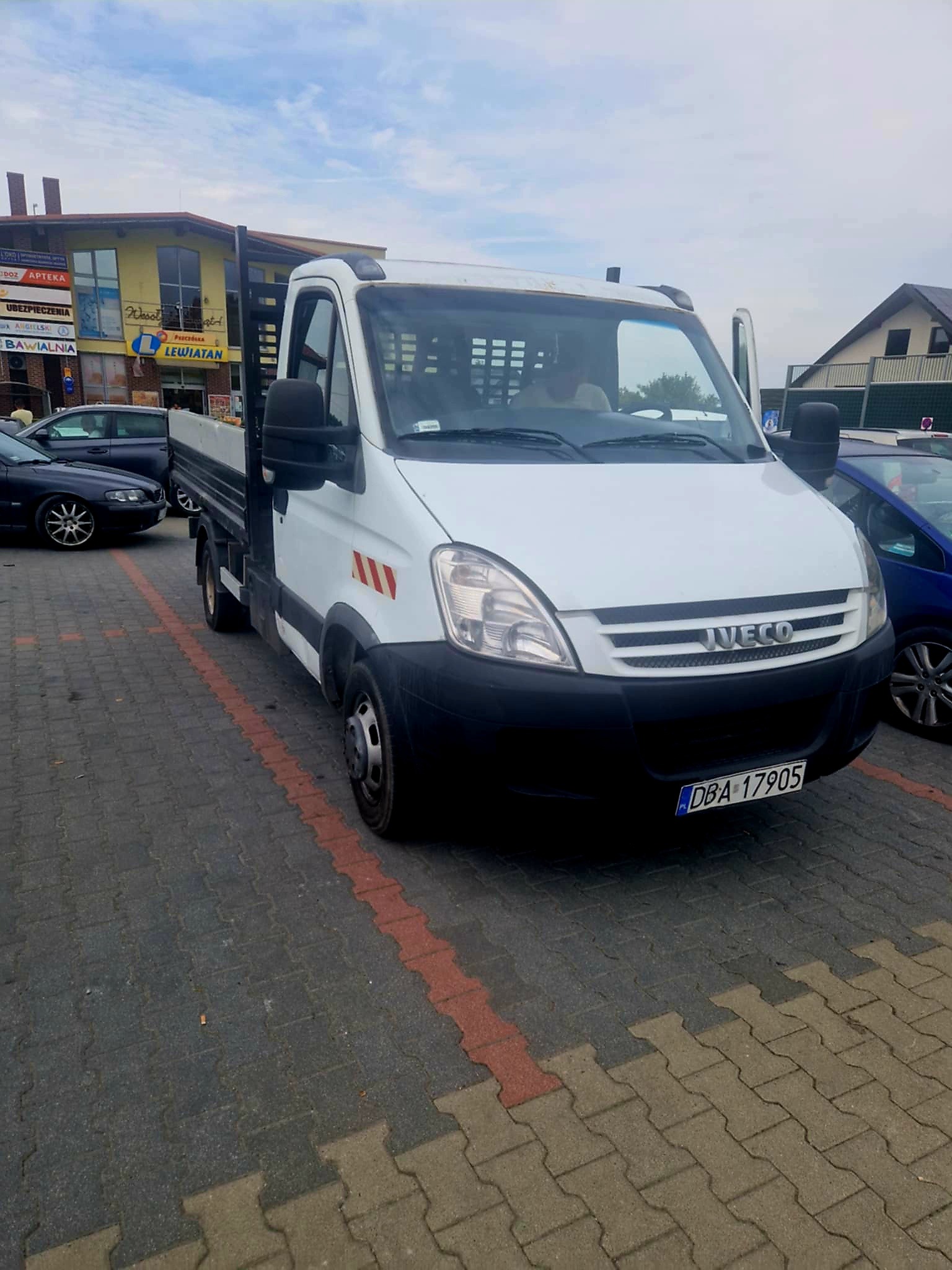 Biały samochód dostawczy marki Iveco z otwartymi drzwiami, zaparkowany na parkingu z kostki brukowej w słoneczny dzień. Widoczny budynek z szyldem 'Lewiatan'.