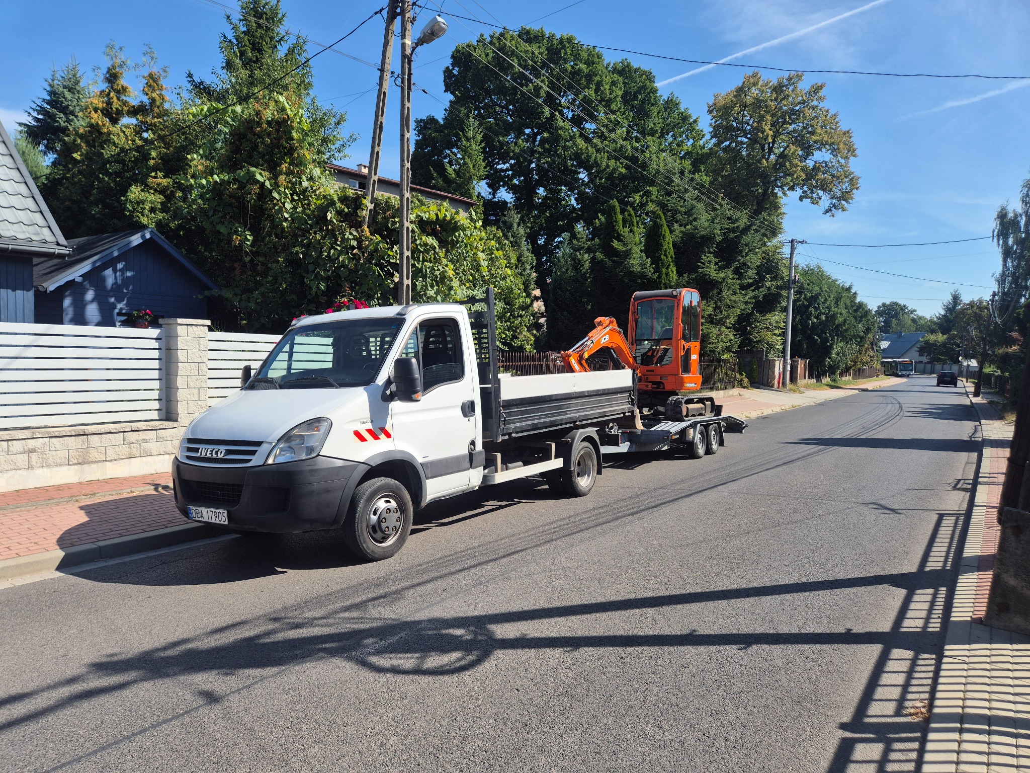 Biały Iveco z przyczepą i pomarańczową mini koparką na ulicy w słoneczny dzień. Widoczne budynki i drzewa w tle. Cień rzucany na asfalt.