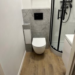 Jarek Dąbrowski - Nowoczesna łazienka po remoncie: podwieszana toaleta, imitacja drewna na podłodze, szara ściana z marmurowym wzorem i czarna armatura w kabinie prysznicowej.