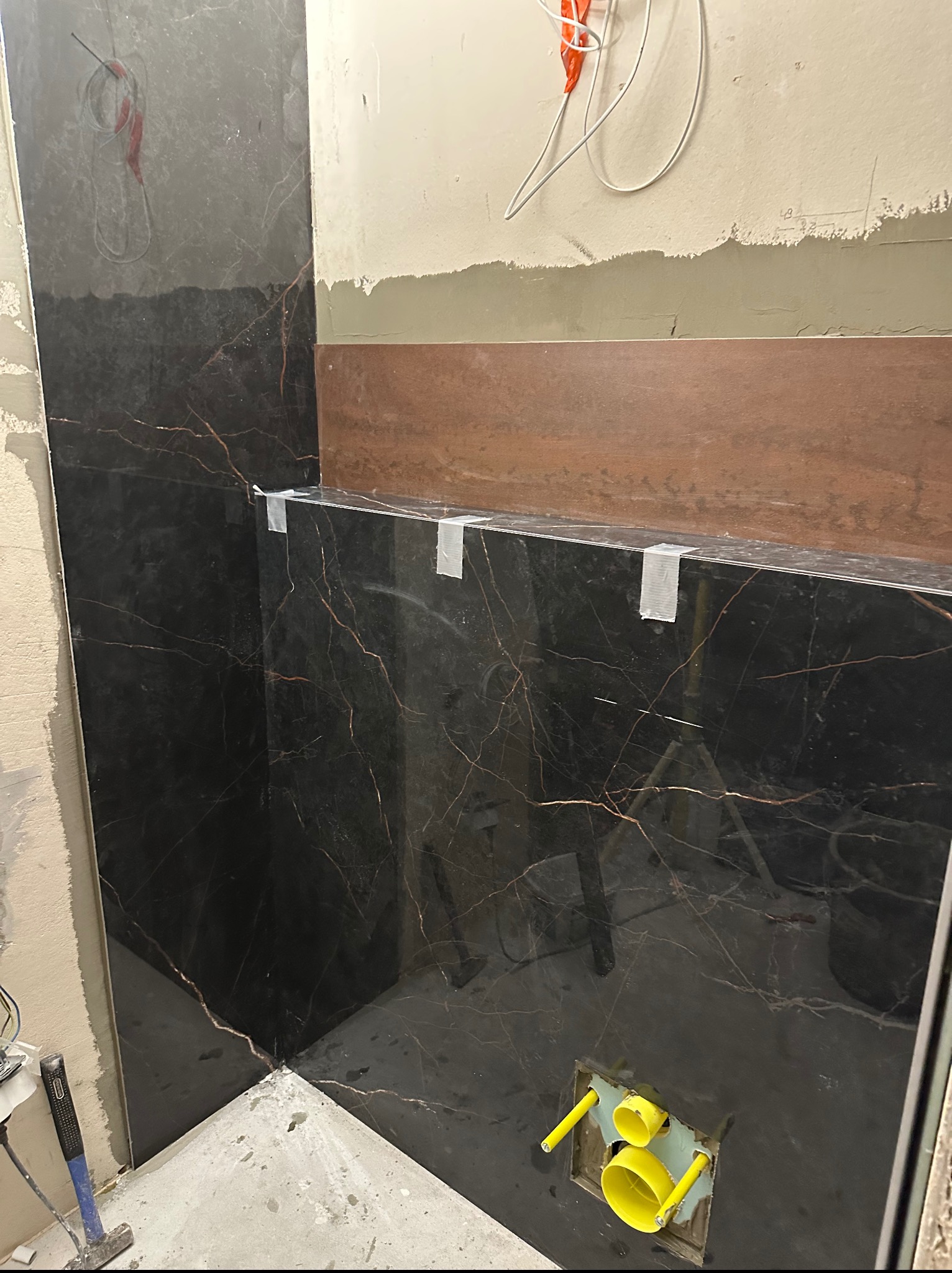 Częściowo wyłożona czarnym marmurem ściana łazienki z widocznymi instalacjami hydraulicznymi, zabezpieczona taśmą malarską, w trakcie prac wykończeniowych.