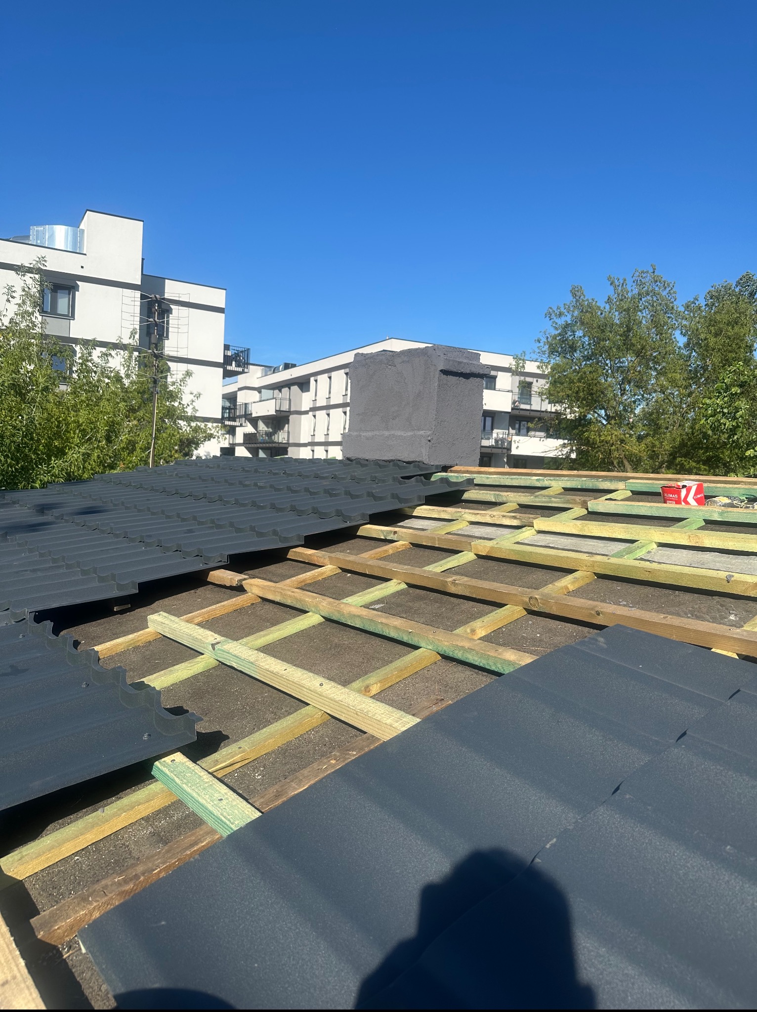 Układanie blachodachówki na drewnianej konstrukcji dachu. Częściowo pokryty dach, w tle budynki mieszkalne i błękitne niebo. Widoczna skrzynka z narzędziami.