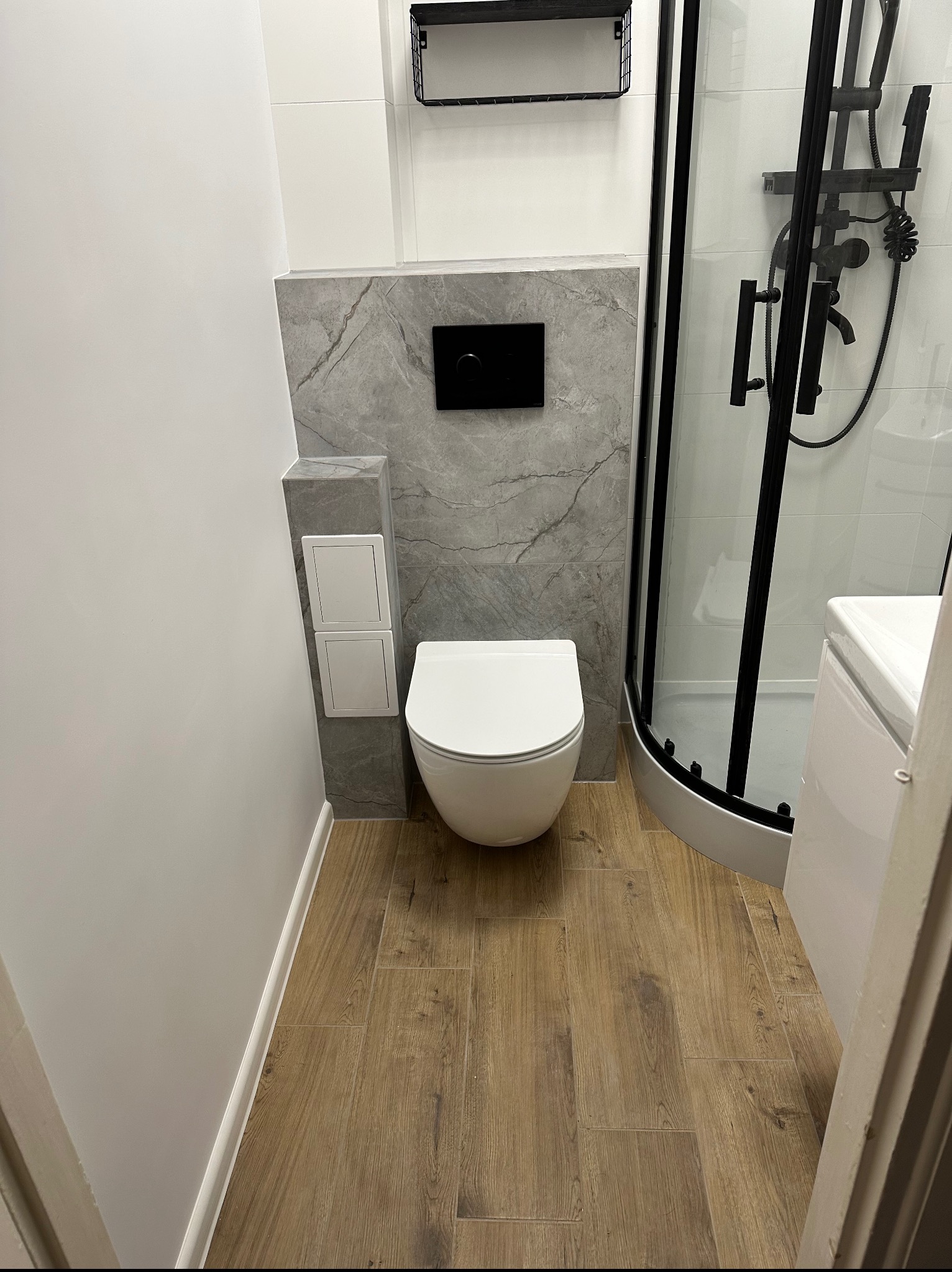 Nowoczesna łazienka po remoncie: podwieszana toaleta, imitacja drewna na podłodze, szara ściana z marmurowym wzorem i czarna armatura w kabinie prysznicowej.