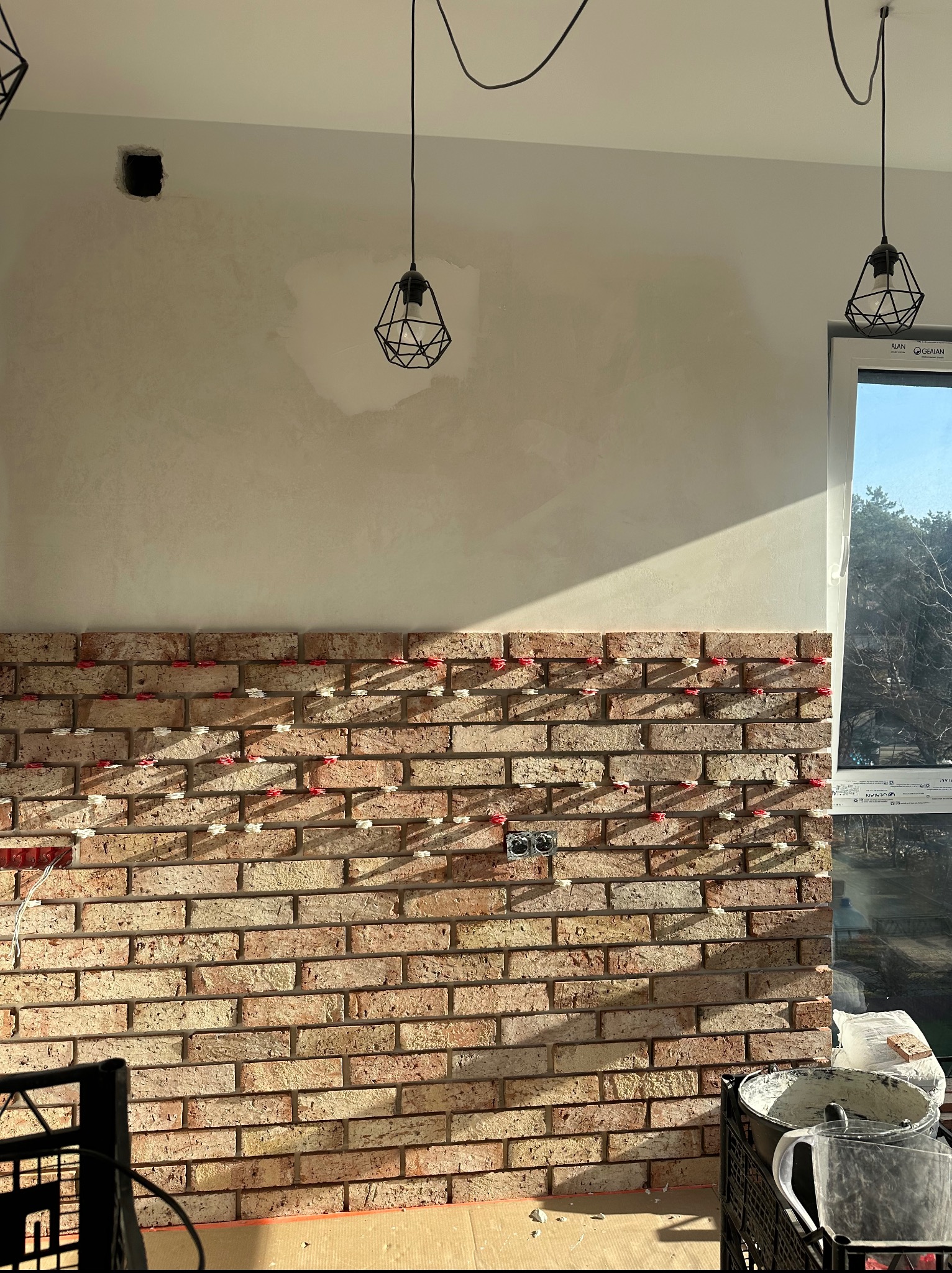 Ściana z cegły klinkierowej w trakcie montażu, widoczne krzyżyki dystansowe i puszka elektryczna. Częściowo wyszpachlowana ściana, okno z widokiem na zewnątrz.