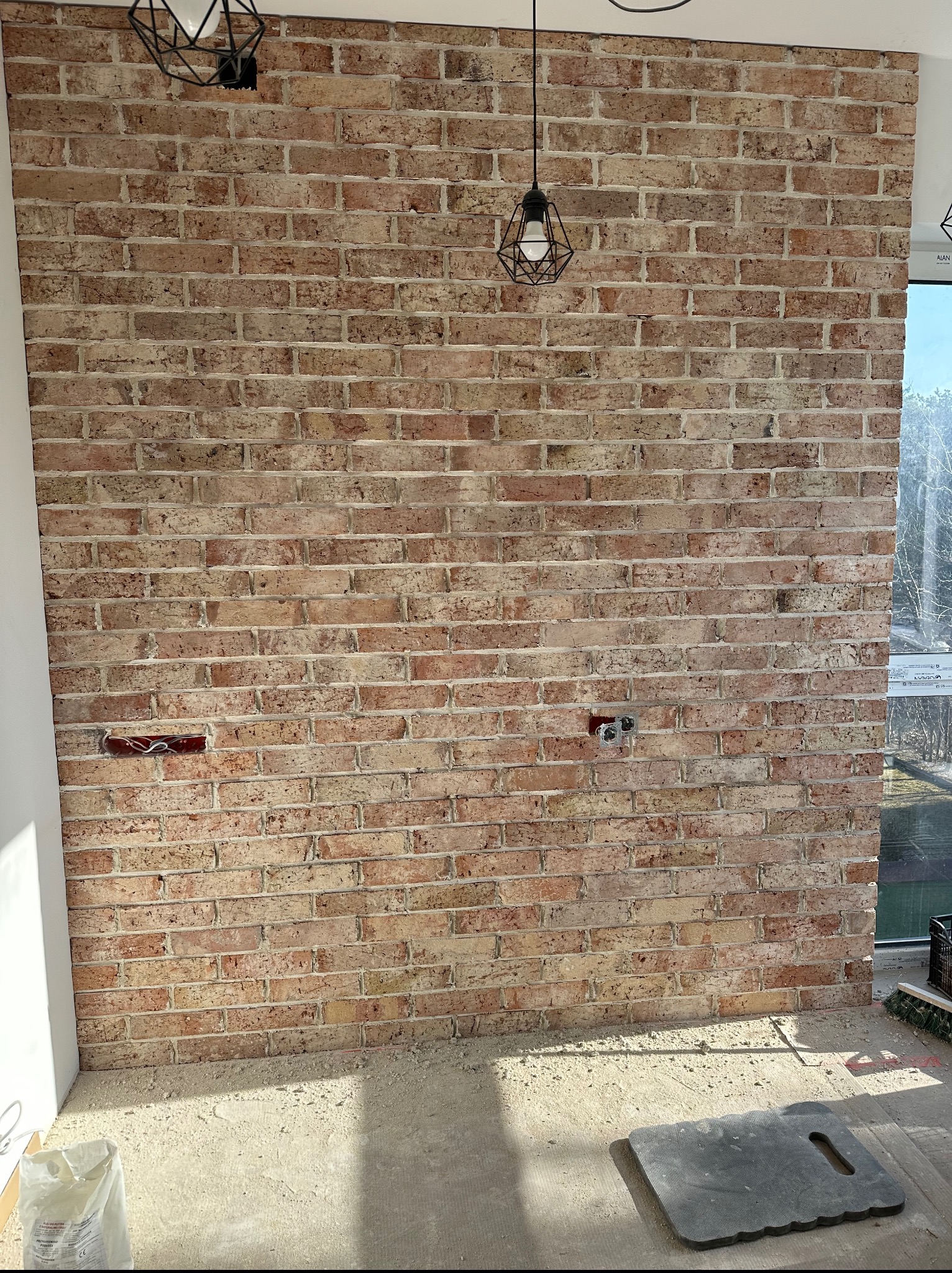 Ściana z cegły klinkierowej z widocznymi gniazdkami elektrycznymi i wiszącymi lampami w geometrycznych oprawach. Widoczne ślady prac wykończeniowych na podłodze.