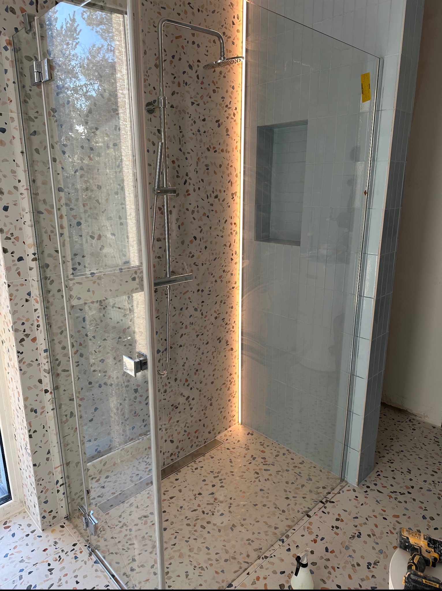 Nowoczesna kabina prysznicowa z deszczownicą i wnęką, wykończona płytkami lastryko i jasnymi, podłużnymi kafelkami. Widoczne narzędzia sugerują prace wykończeniowe.