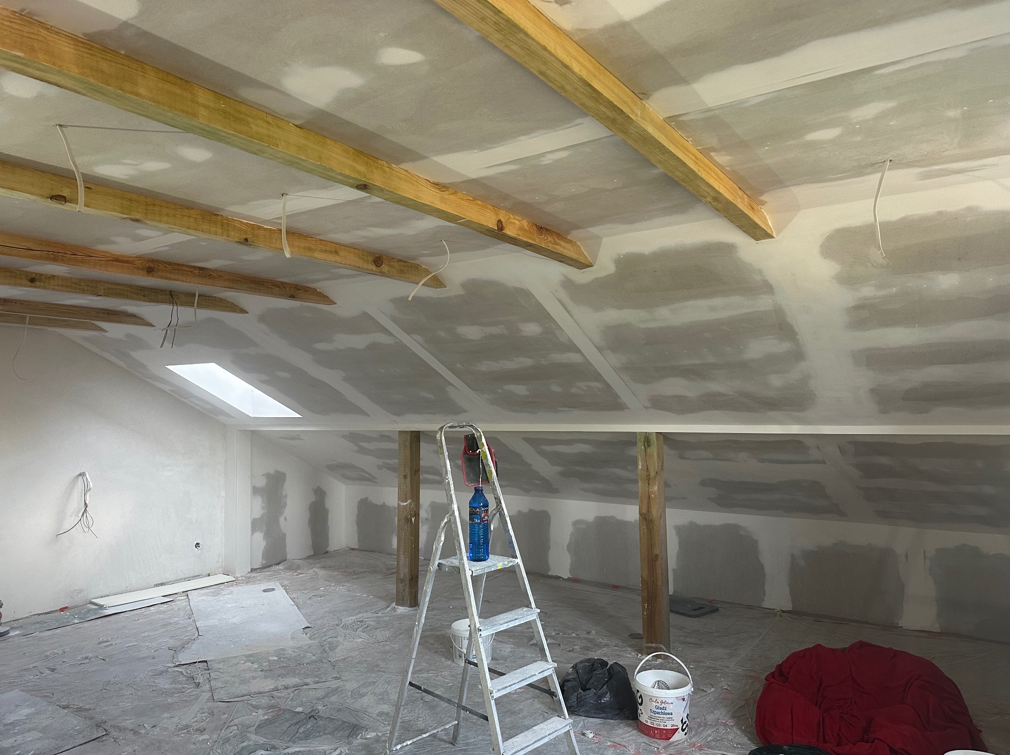 Poddasze w trakcie prac wykończeniowych: widoczne drewniane belki stropowe, płyty gipsowo-kartonowe, szpachlowanie, drabina i akcesoria malarskie. Skos dachu z oknem.
