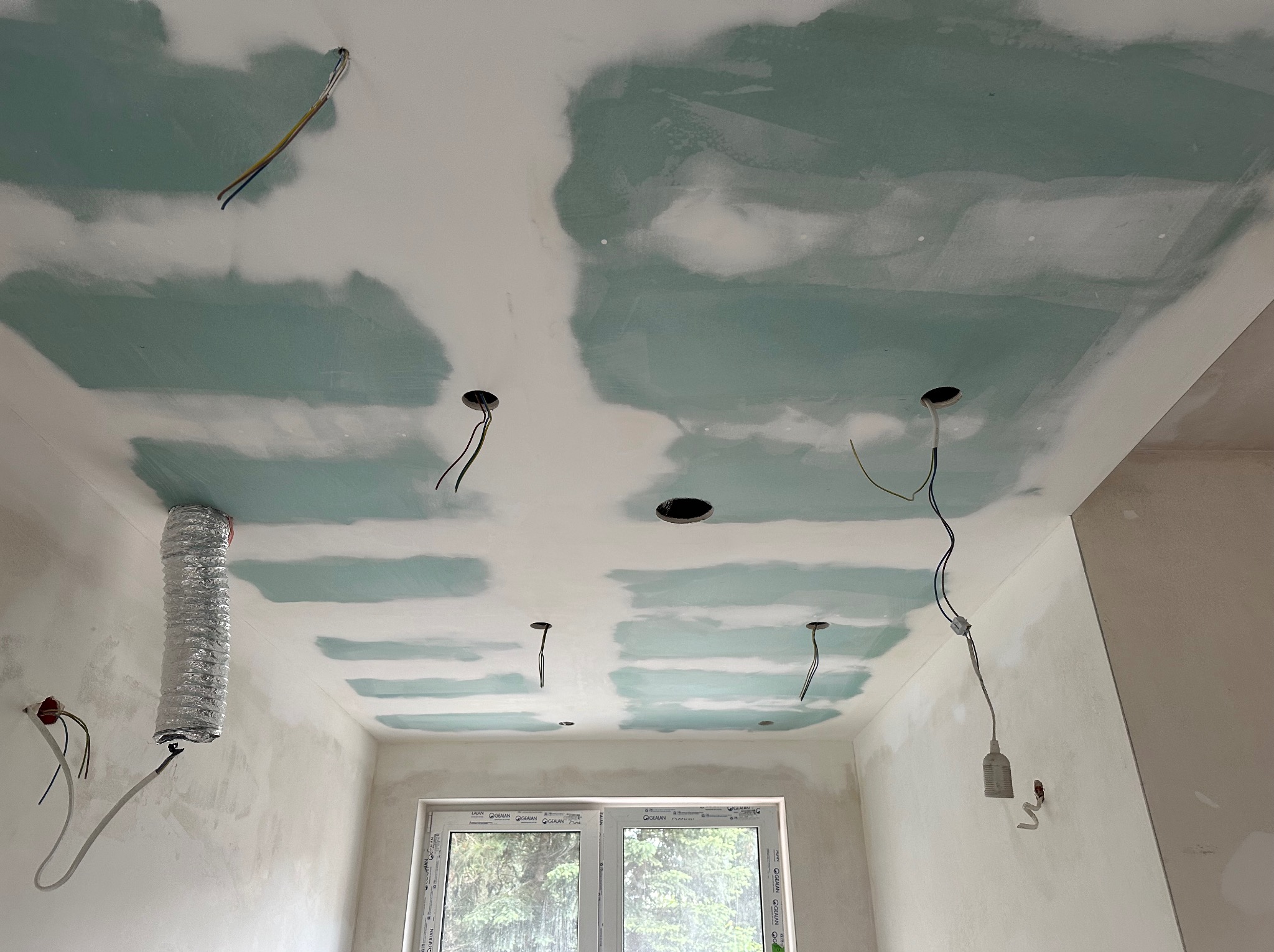 Sufit z płyt gipsowo-kartonowych w trakcie szpachlowania, widoczne otwory na oświetlenie i wystające przewody, wentylacja. Ściany przygotowane do malowania.