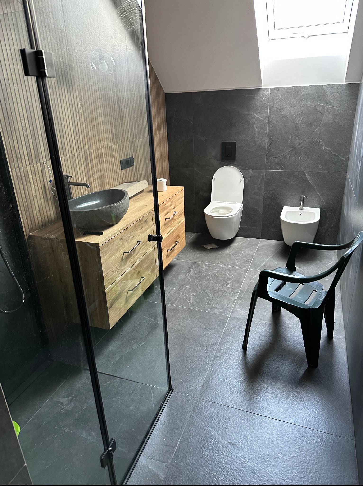 Nowoczesna łazienka z kabiną prysznicową, drewnianą szafką z kamienną umywalką i szarą armaturą. Uwagę zwraca okno dachowe w skosie.