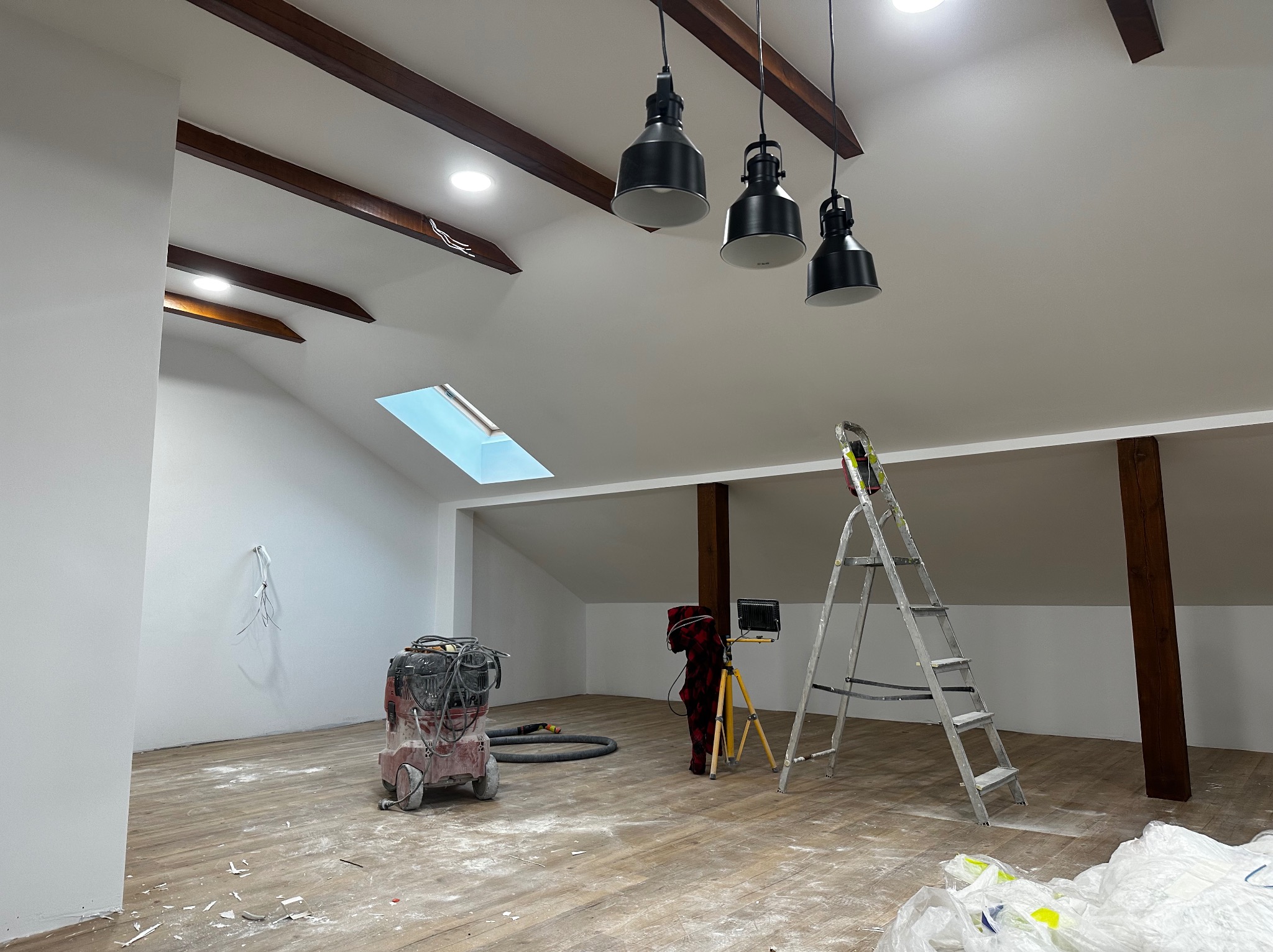 Poddasze po szpachlowaniu ścian, z widocznymi belkami stropowymi, oknem dachowym, lampami i narzędziami: odkurzacz przemysłowy i drabina. Podłoga częściowo zakryta folią.