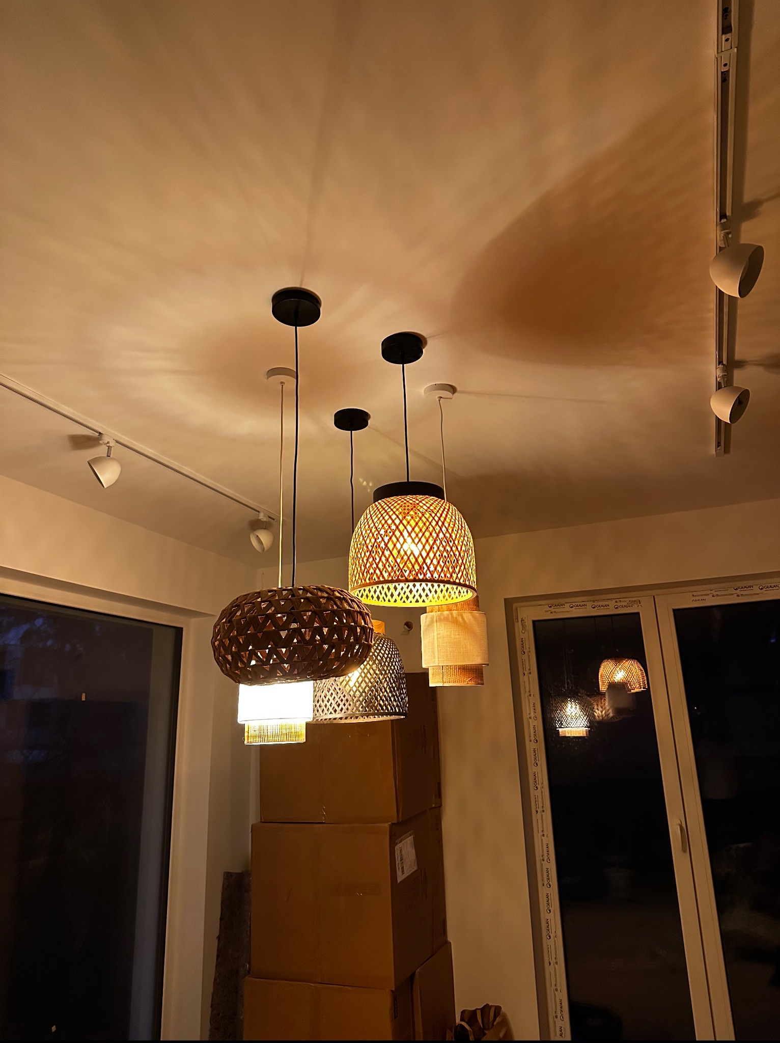 Cztery lampy wiszące o różnych wzorach i materiałach, oświetlające pomieszczenie z widocznymi kartonami i oknami. Dodatkowe oświetlenie punktowe na suficie.