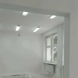 Tomasz Woroniecki Profesjonalne technologie natryskowe w budownictwie i przemyśle - Białe, odświeżone wnętrze po remoncie. Widok na ścianę z oknem i grzejnikiem, oświetloną przez podłużne lampy sufitowe. Na podłodze widoczne resztki folii malarskiej.