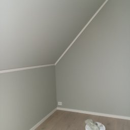 Tomasz Woroniecki Profesjonalne technologie natryskowe w budownictwie i przemyśle - Pomieszczenie po odświeżeniu ścian szarą farbą. Widoczny skośny sufit, listwy przypodłogowe i wiadro z farbą. Gładkie ściany w odcieniach szarości.