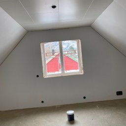 Tomasz Woroniecki Profesjonalne technologie natryskowe w budownictwie i przemyśle - Poddasze z oknem i puszkami elektrycznymi przed malowaniem. Na podłodze stoi puszka z farbą. Widok na czerwony dom za oknem. Ściany przygotowane do malowania.