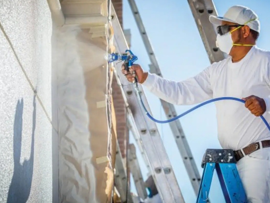 Pracownik w masce i okularach natryskuje farbę na elewację budynku z użyciem pistoletu natryskowego. Widoczna drabina i fragment pomalowanej ściany w słoneczny dzień.