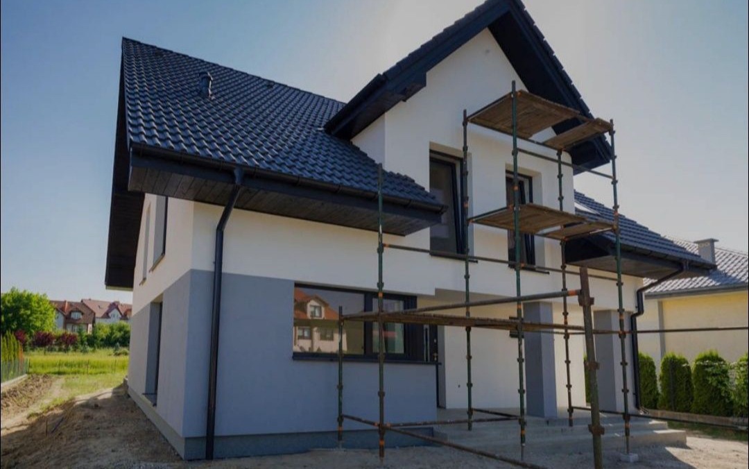 Dwukolorowa elewacja domu jednorodzinnego w trakcie malowania, z widocznymi rusztowaniami. Dolna część szara, górna biała, czarny dach i okna.