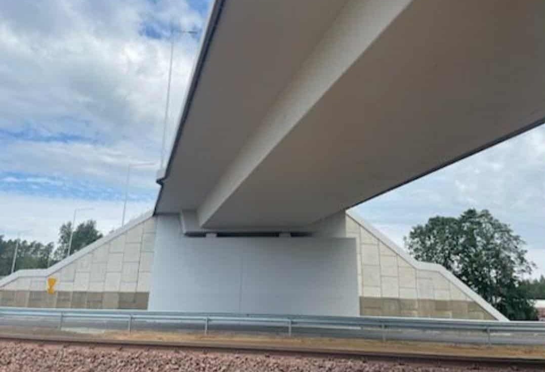 Jasny, betonowy wiadukt z widoczną konstrukcją nośną i fragmentem nieba w tle. Widoczne elementy infrastruktury drogowej i fragment zieleni.