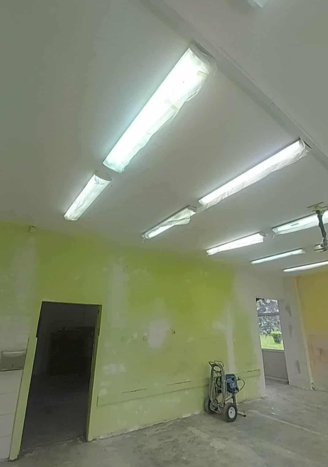 Wnętrze w trakcie prac przygotowawczych: zabezpieczone lampy sufitowe, ściany z widocznymi śladami po szpachlowaniu i resztkach farby, agregat malarski w rogu.