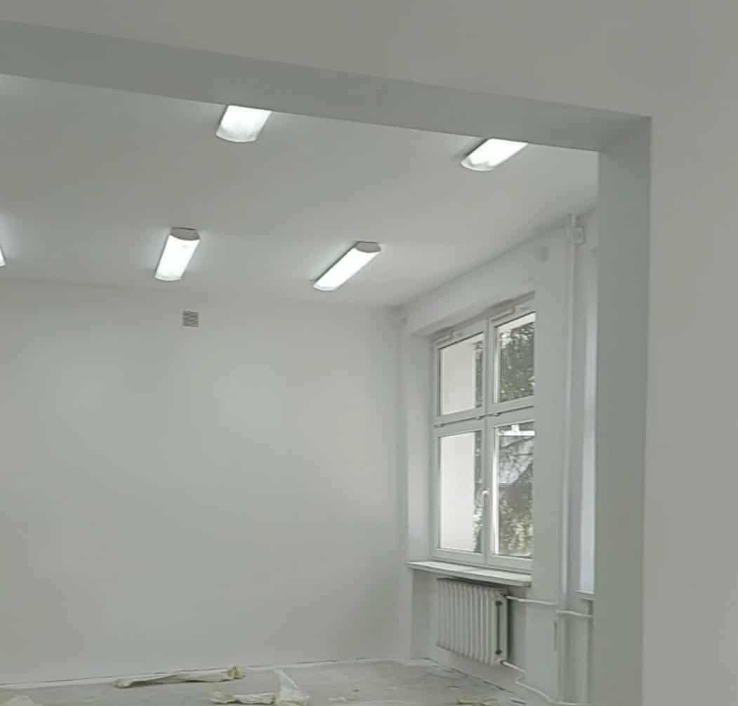 Białe, odświeżone wnętrze po remoncie. Widok na ścianę z oknem i grzejnikiem, oświetloną przez podłużne lampy sufitowe. Na podłodze widoczne resztki folii malarskiej.