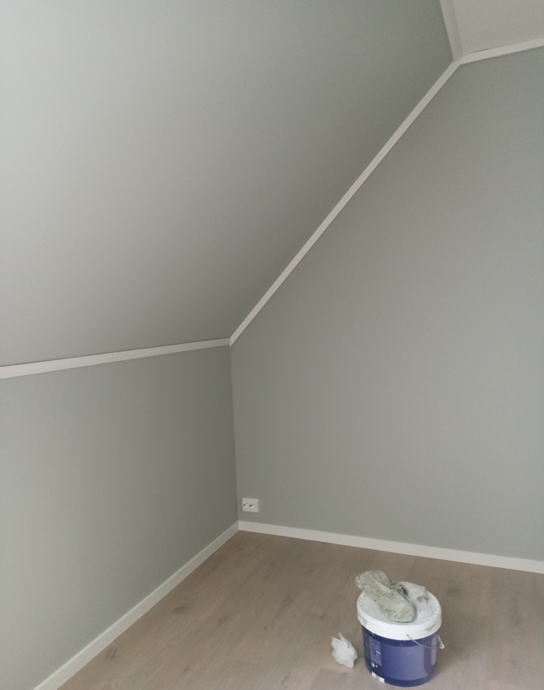Pomieszczenie po odświeżeniu ścian szarą farbą. Widoczny skośny sufit, listwy przypodłogowe i wiadro z farbą. Gładkie ściany w odcieniach szarości.