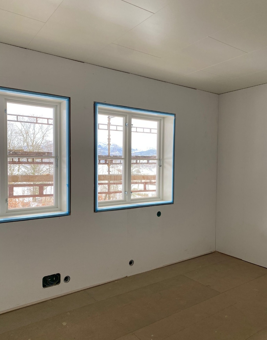 Pokój po szpachlowaniu, przygotowany do malowania. Okna zabezpieczone taśmą, widoczne puszki elektryczne w ścianie. Podłoga zabezpieczona kartonem.