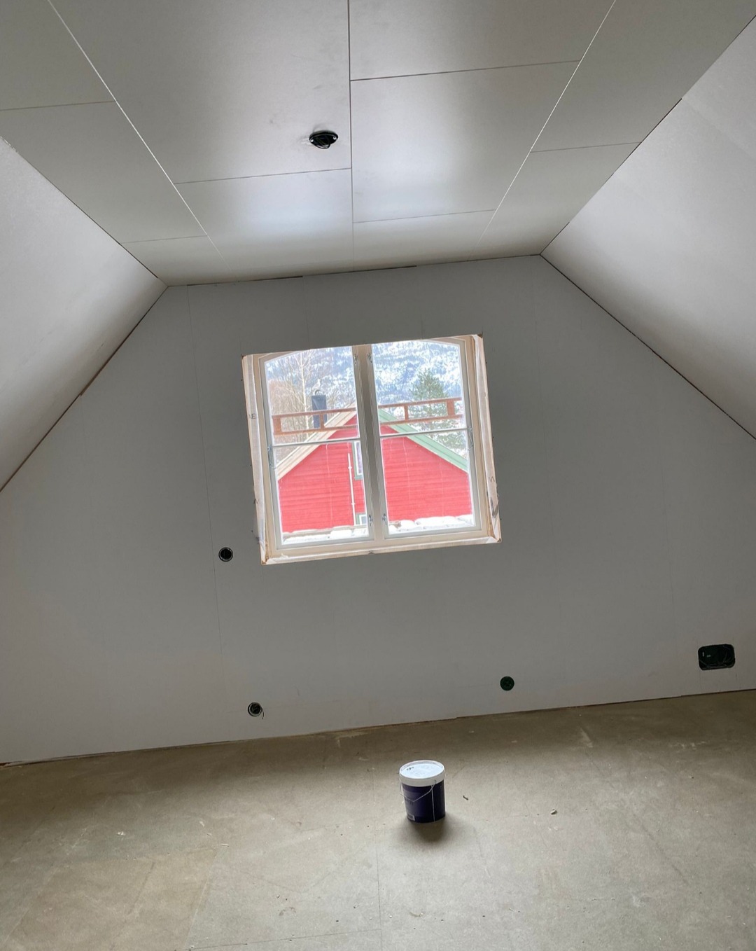 Poddasze z oknem i puszkami elektrycznymi przed malowaniem. Na podłodze stoi puszka z farbą. Widok na czerwony dom za oknem. Ściany przygotowane do malowania.