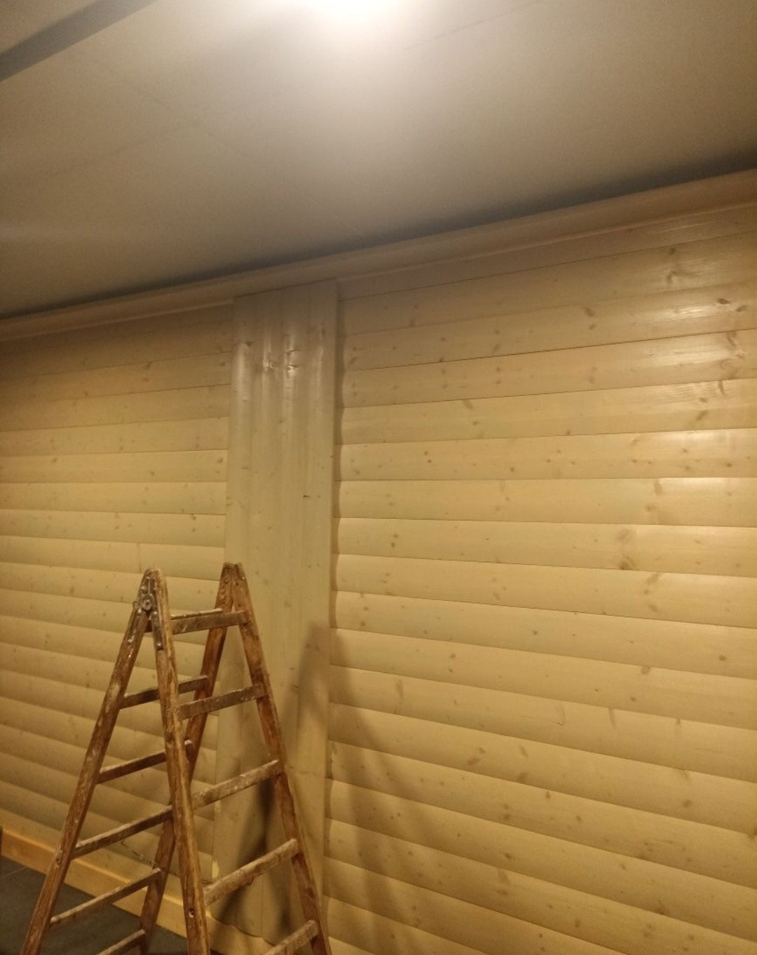 Drewniana ściana z boazerią, częściowo zabezpieczona folią malarską, z drewnianą drabiną opartą o ścianę. Przygotowanie do prac wykończeniowych.