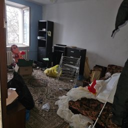 WYWÓZ PRO - Opróżnianie mieszkań I wywóz mebli - Pokój przed opróżnianiem: zagracony dywan, meble, kartony, dmuchany bokser przy oknie. Widok na zaniedbane wnętrze przed usługą porządkową.