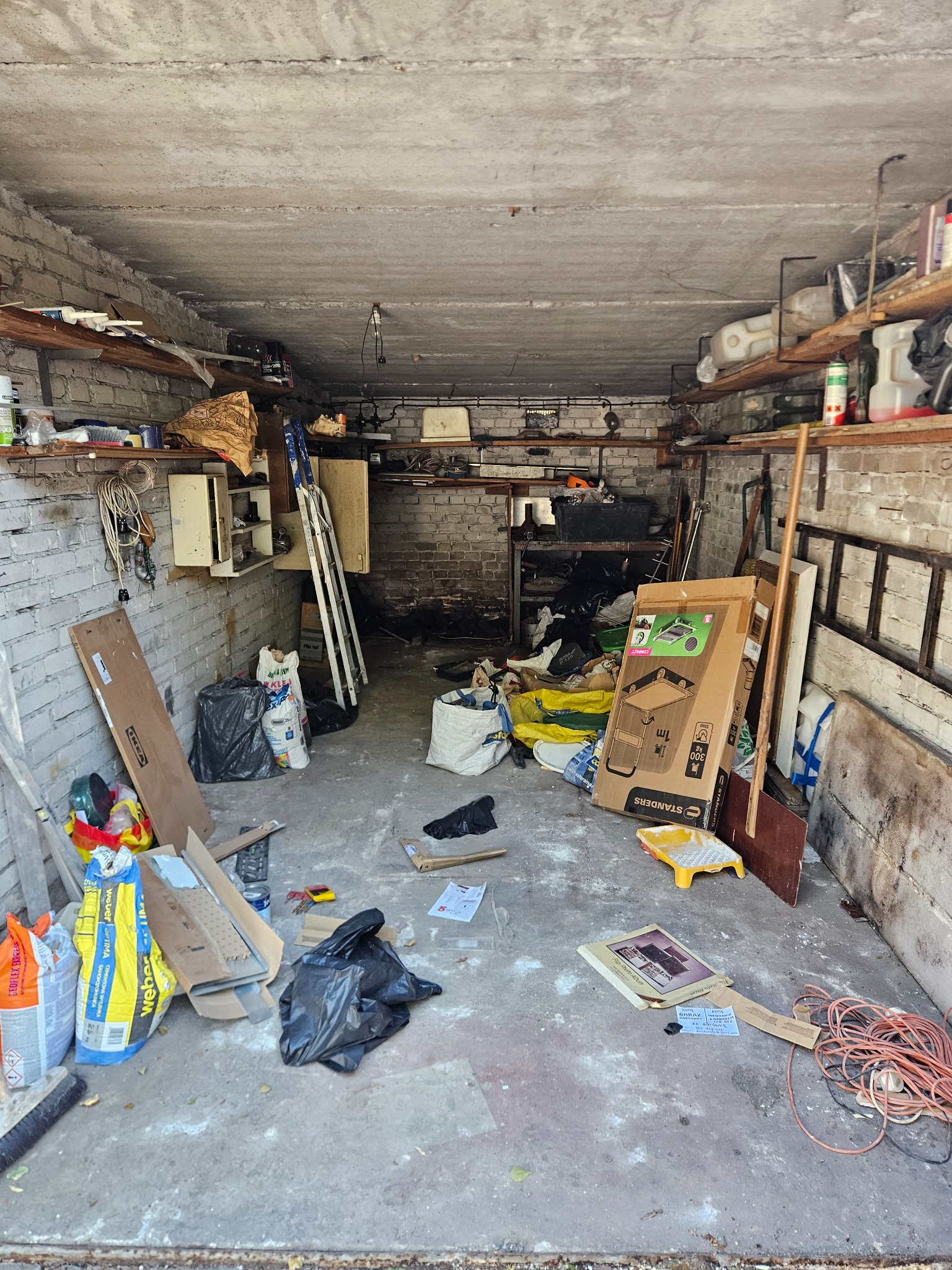 Zagracony garaż z ceglanymi ścianami i betonową podłogą. Widoczne worki, kartony, narzędzia i inne przedmioty. Panuje nieporządek, co sugeruje potrzebę opróżnienia.