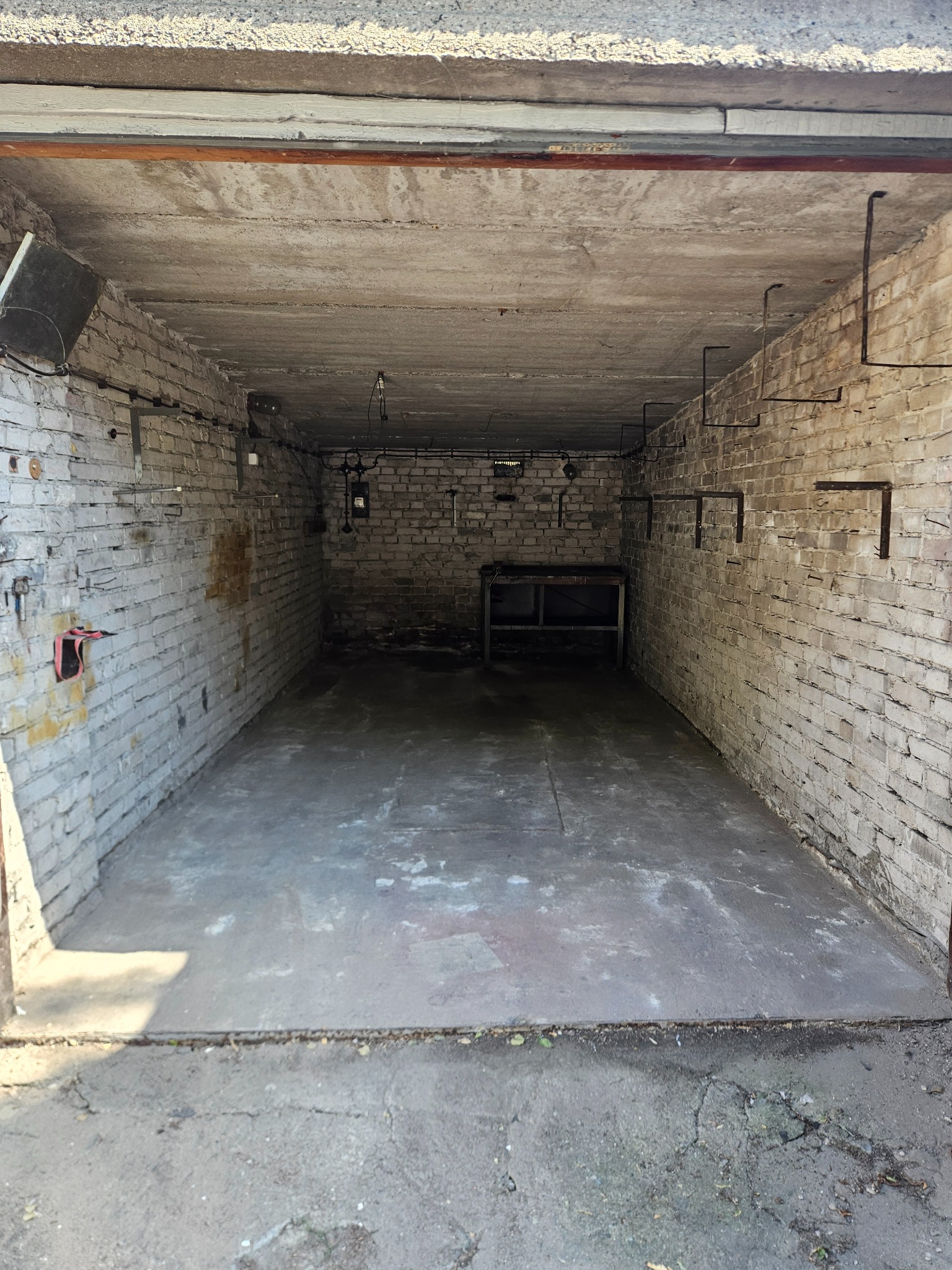 Opróżniony garaż z białymi ceglanymi ścianami i betonową podłogą. W tle widoczny stół warsztatowy. Widok z zewnątrz, światło dzienne.