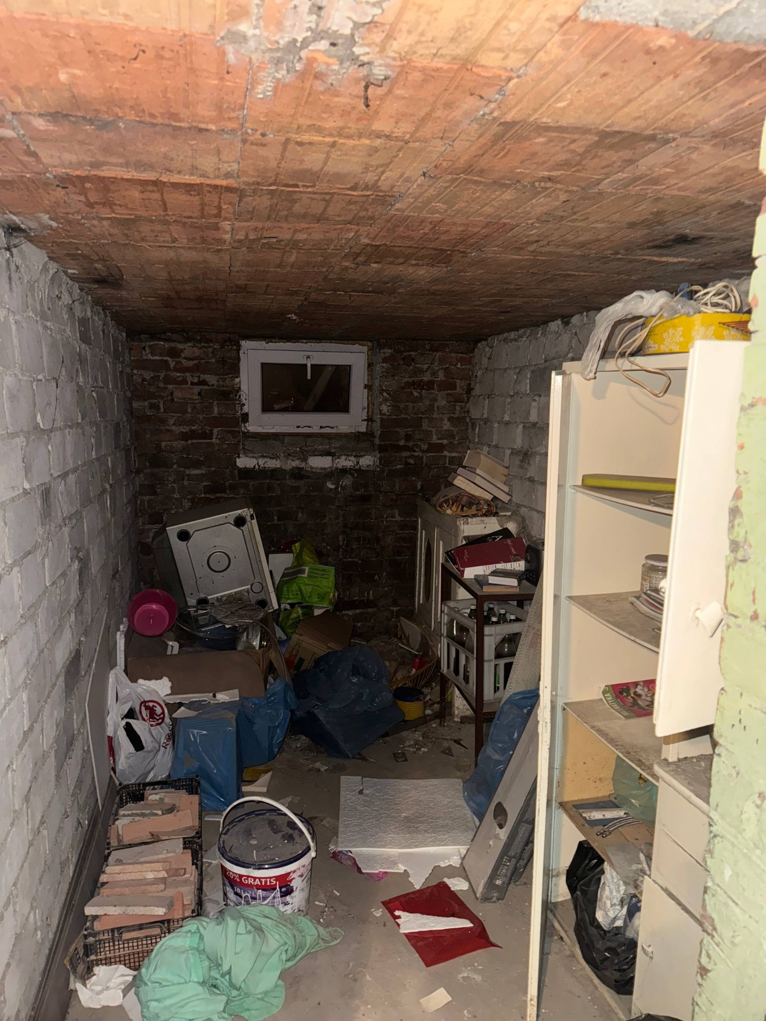 Zagracona piwnica z ceglanym sufitem, widoczne stare meble, worki na śmieci, cegły i wiadro z farbą. Nierówna podłoga i ściany. Małe okno w tle.