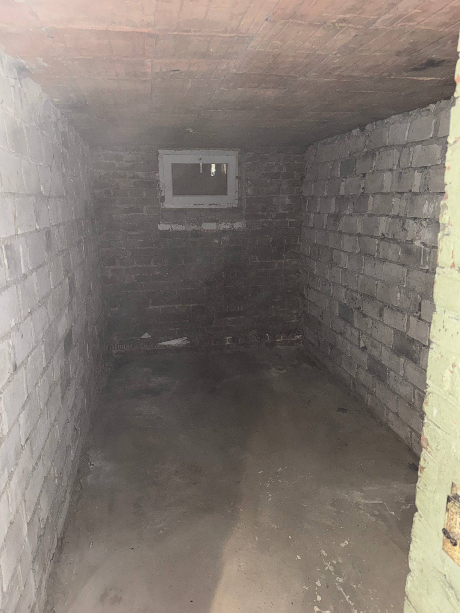 Pusta piwnica z ceglanymi ścianami i małym oknem. Podłoga betonowa, surowy stan. Widok na wnętrze po opróżnianiu, ukazujący czystą przestrzeń.