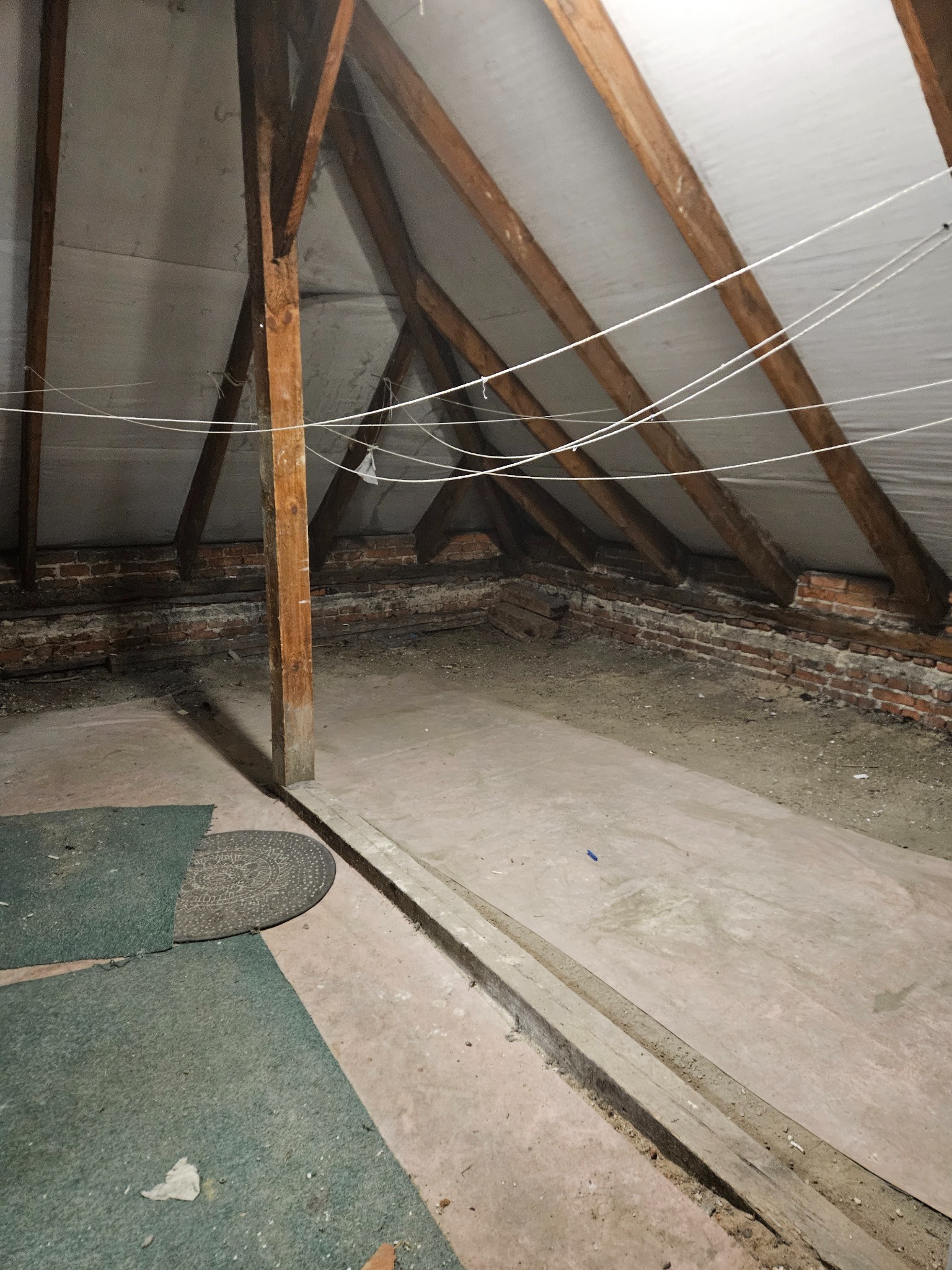 Pusty strych z drewnianą konstrukcją dachu, ceglaną ścianką i resztkami gruzu na podłodze. Widoczne stare dywany i wystające kable.