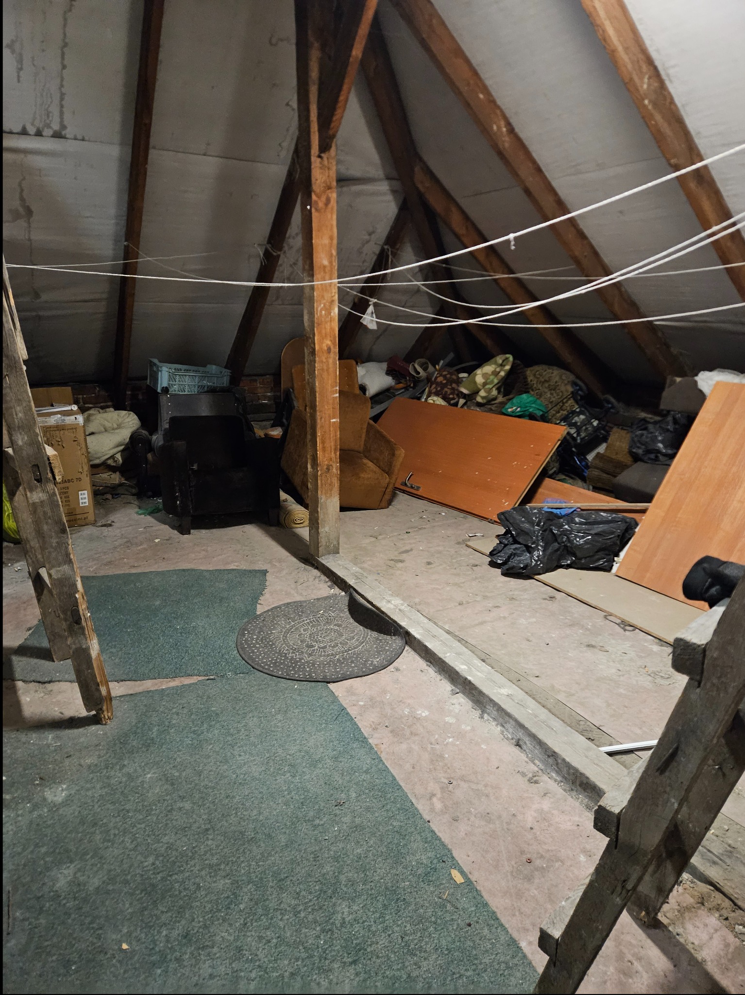 Zagracony strych przed opróżnianiem: stare meble, kartony, dywany, worki na śmieci. Widoczne drewniane elementy konstrukcyjne dachu i drabina. Nierówna podłoga.