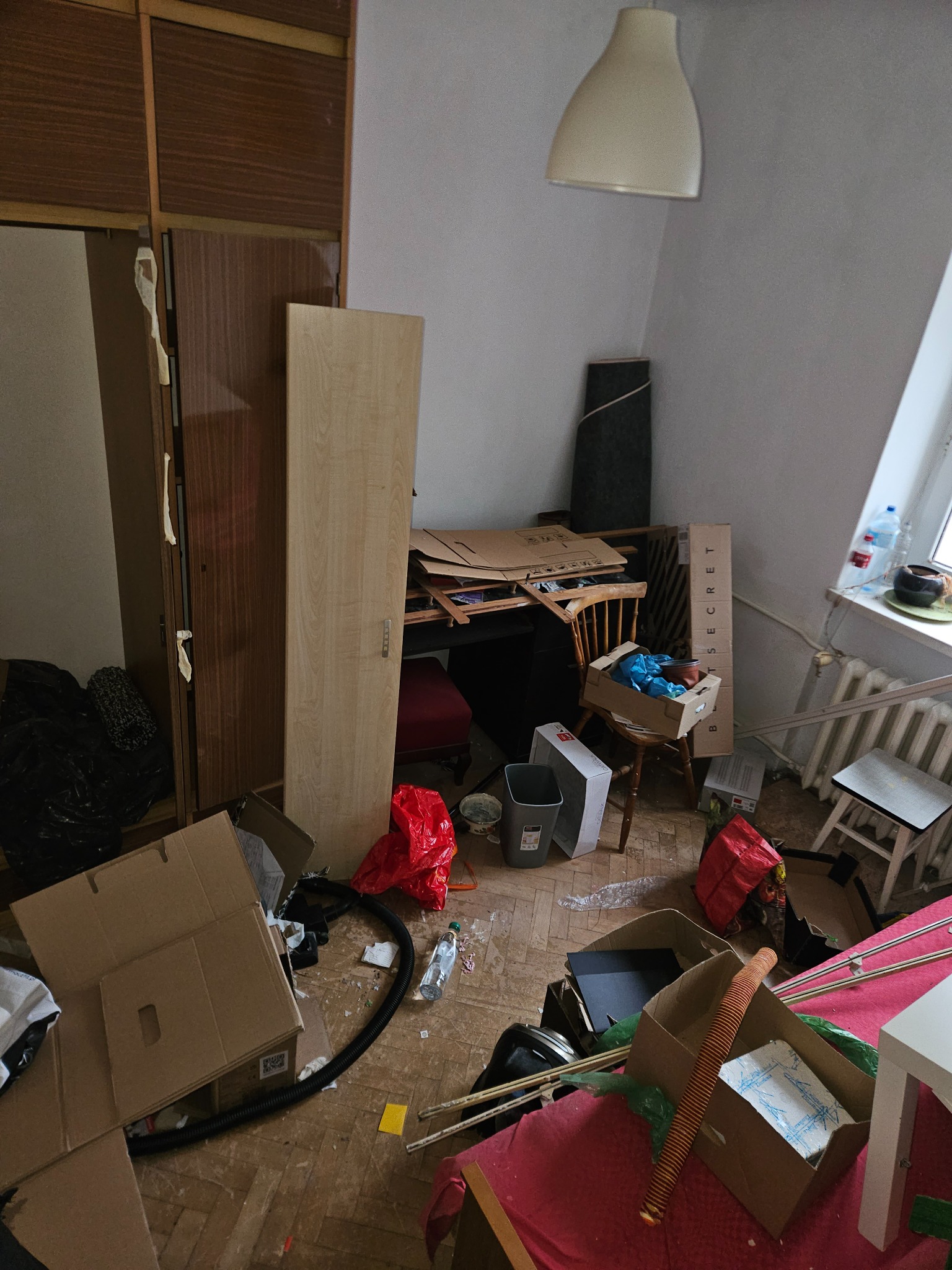 Pokój zagracony kartonami, meblami i śmieciami. Widok na podłogę z parkietem, szafę, biurko i okno. Scena przed opróżnianiem mieszkania.