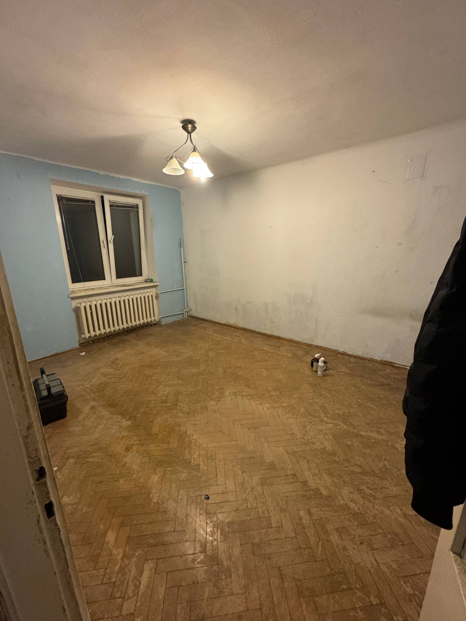 Pusty pokój po opróżnianiu: parkiet w jodełkę, ściany z plamami, okno z grzejnikiem, lampa sufitowa. W rogu skrzynka narzędziowa i kurtka. Efekt po usunięciu zawartości.