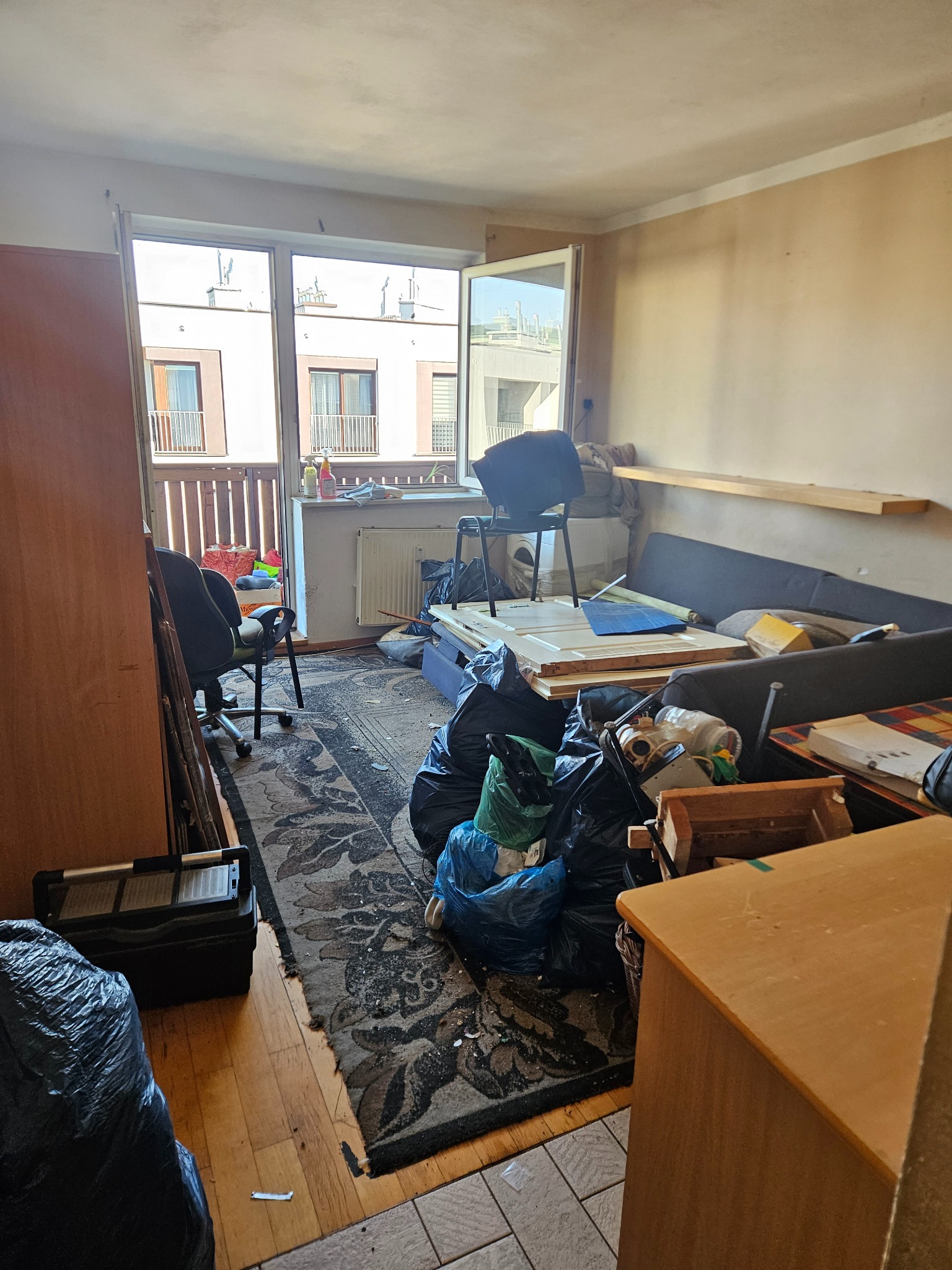 Zabałaganione wnętrze pokoju przed opróżnianiem: worki na śmieci, meble, dywan. Widok na balkon i budynek naprzeciwko. Scena przed rozpoczęciem prac porządkowych.